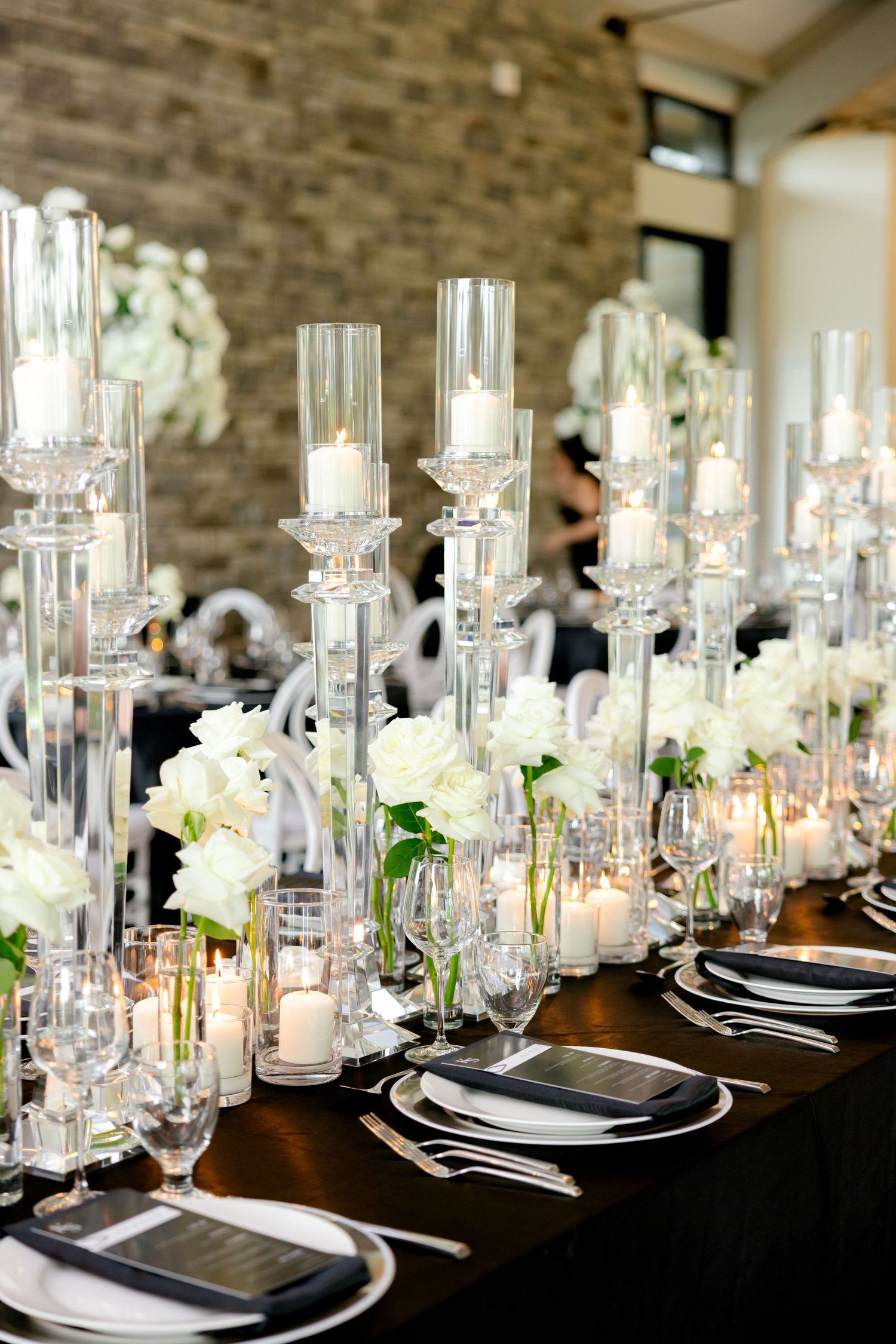 Elegant wedding table setting with tall glass candleholders and white floral arrangements on dark tablecloth.