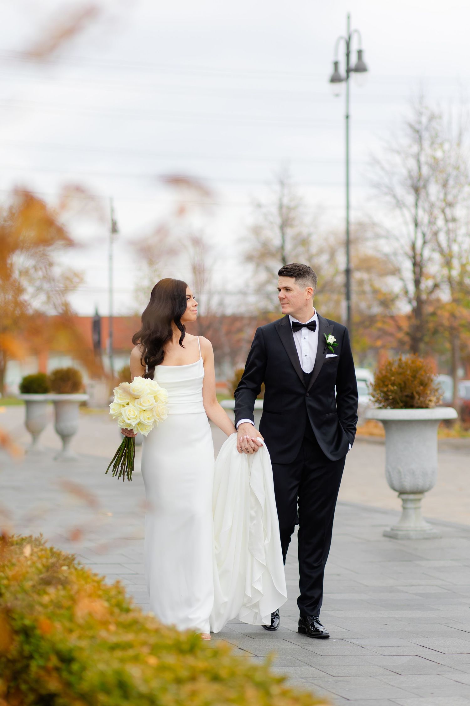 A wedding couple walks together on a stone patio surrounded by autumn foliage and decorative stone urns.