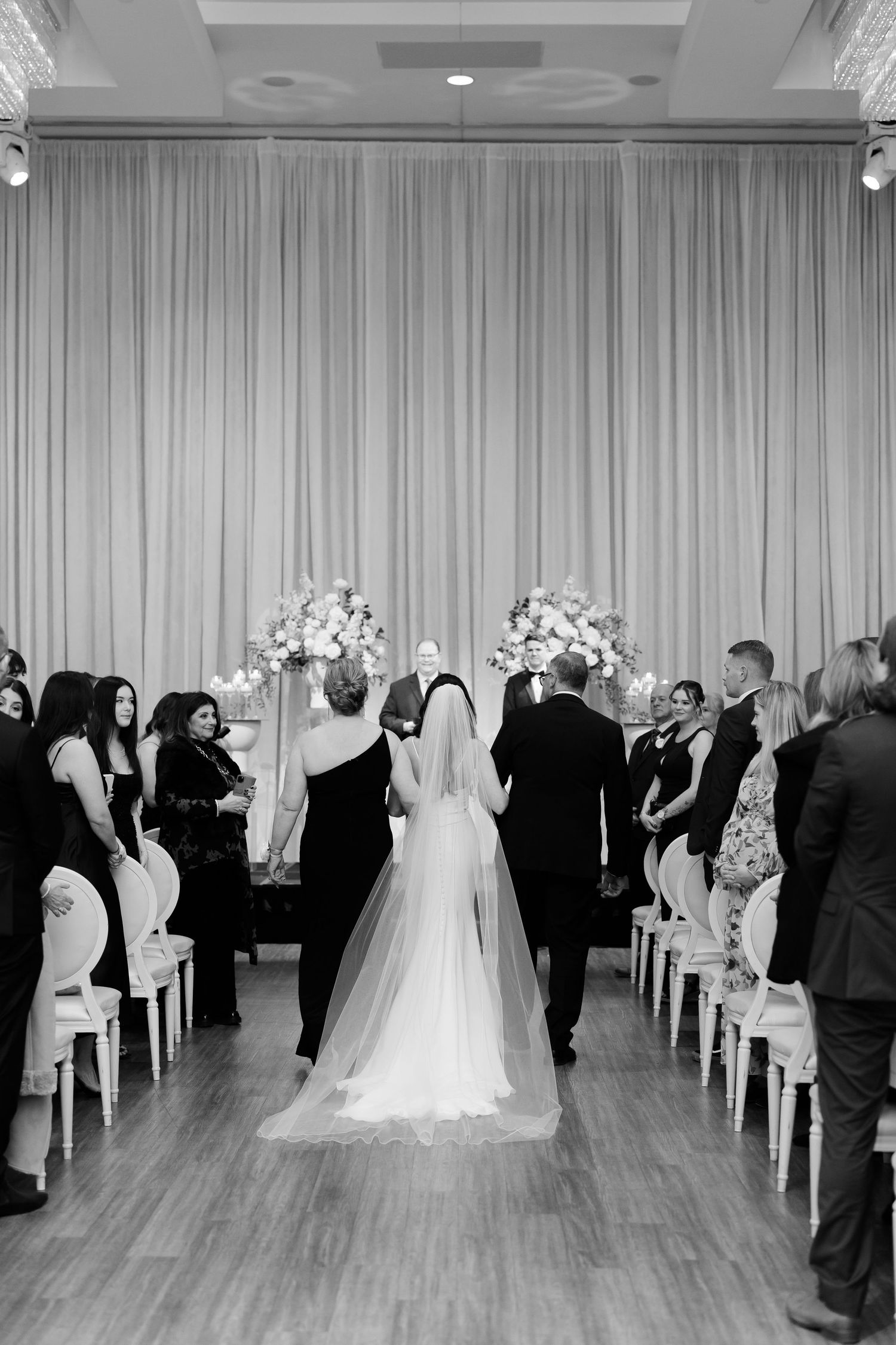 Wedding ceremony in progress with guests seated in white chairs and floral arrangements at the altar.