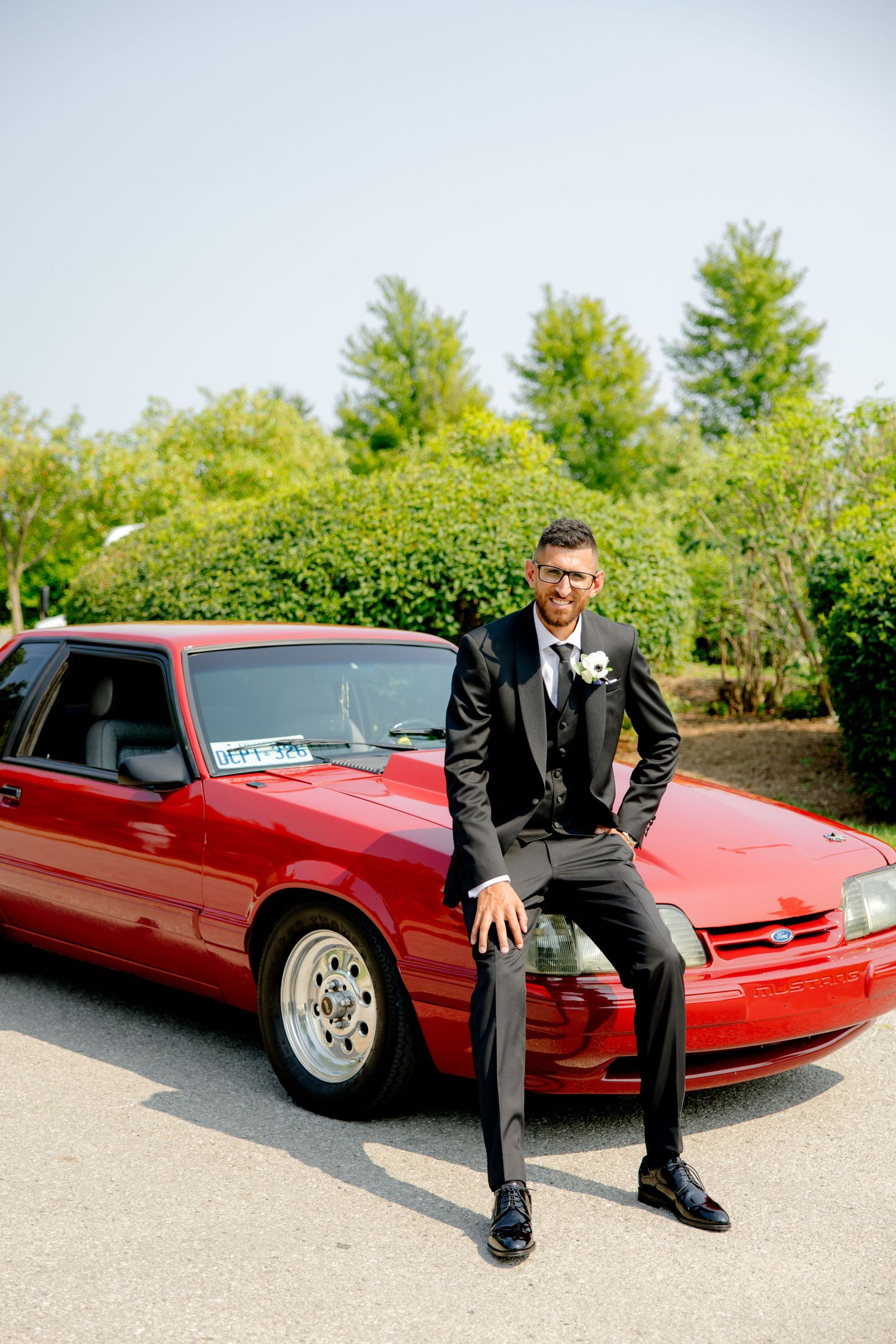 Groom in black tuxedo sits on hood of red sports car with trees in background.