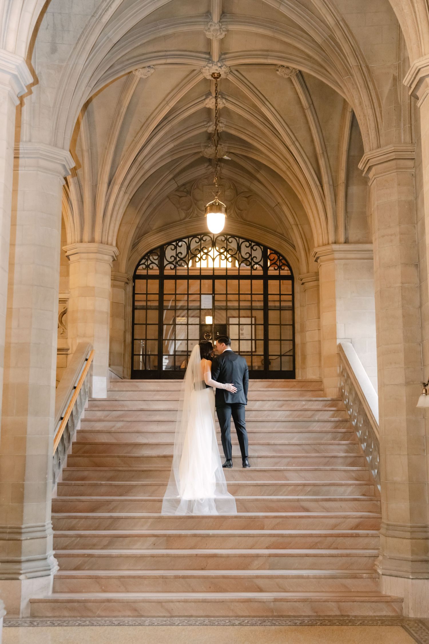 Newlyweds ascend elegant stone staircase under Gothic arches in historic building.
