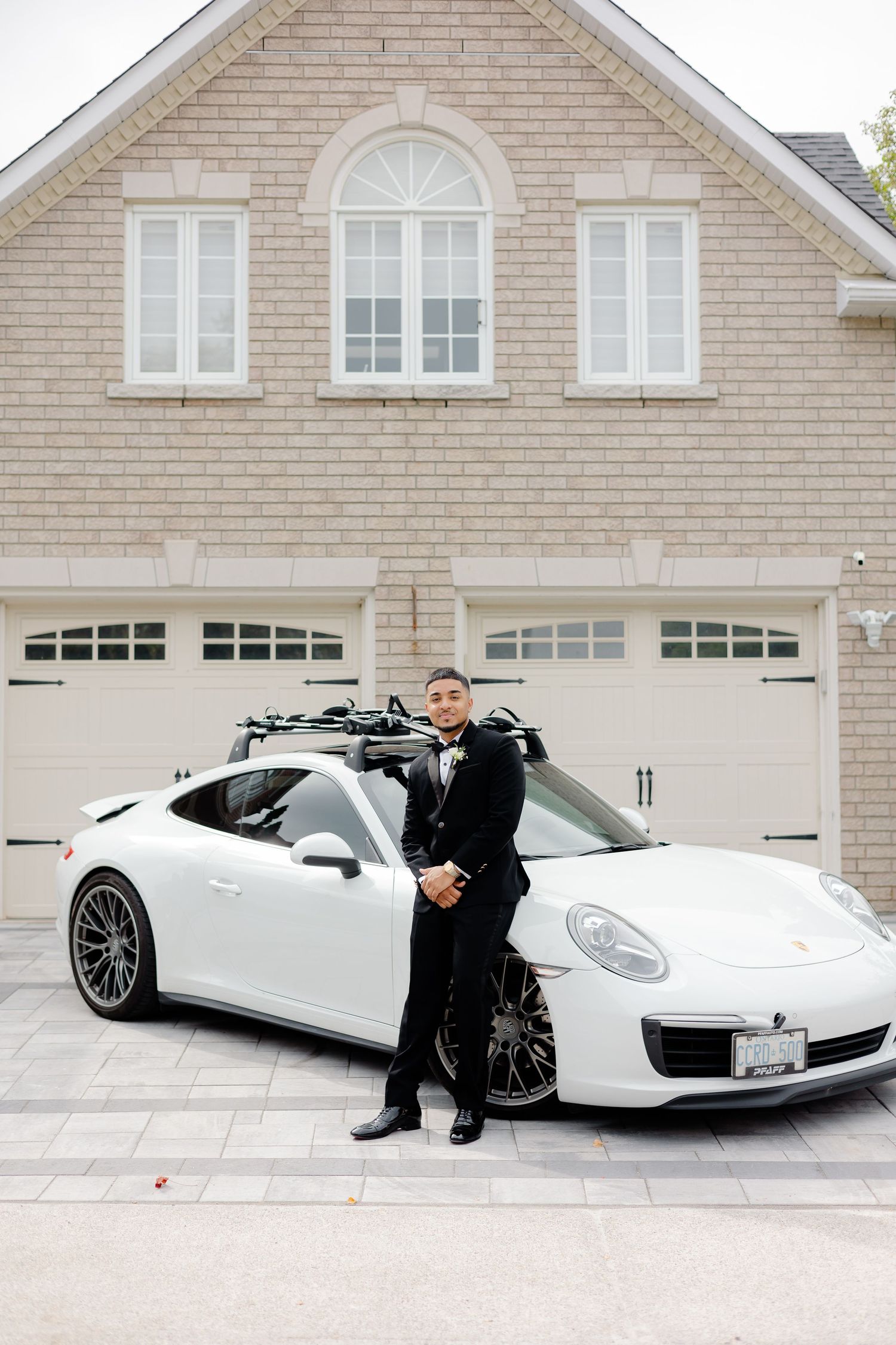 Someone in a black tuxedo poses in front of a white Porsche and beige brick house with double garage.