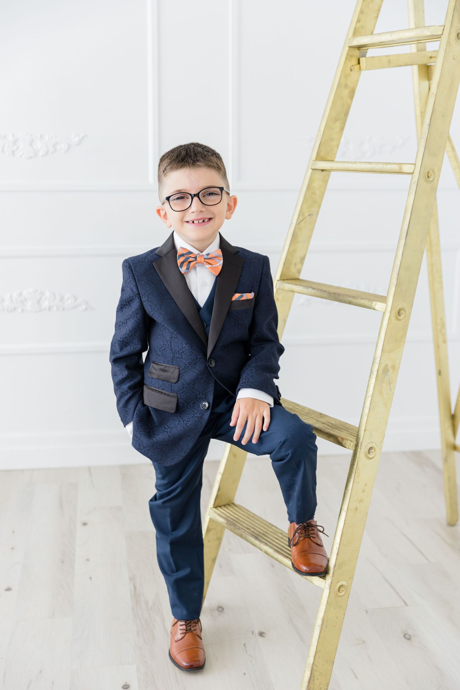 Youth in formal attire poses with foot on gold ladder against white wall.
