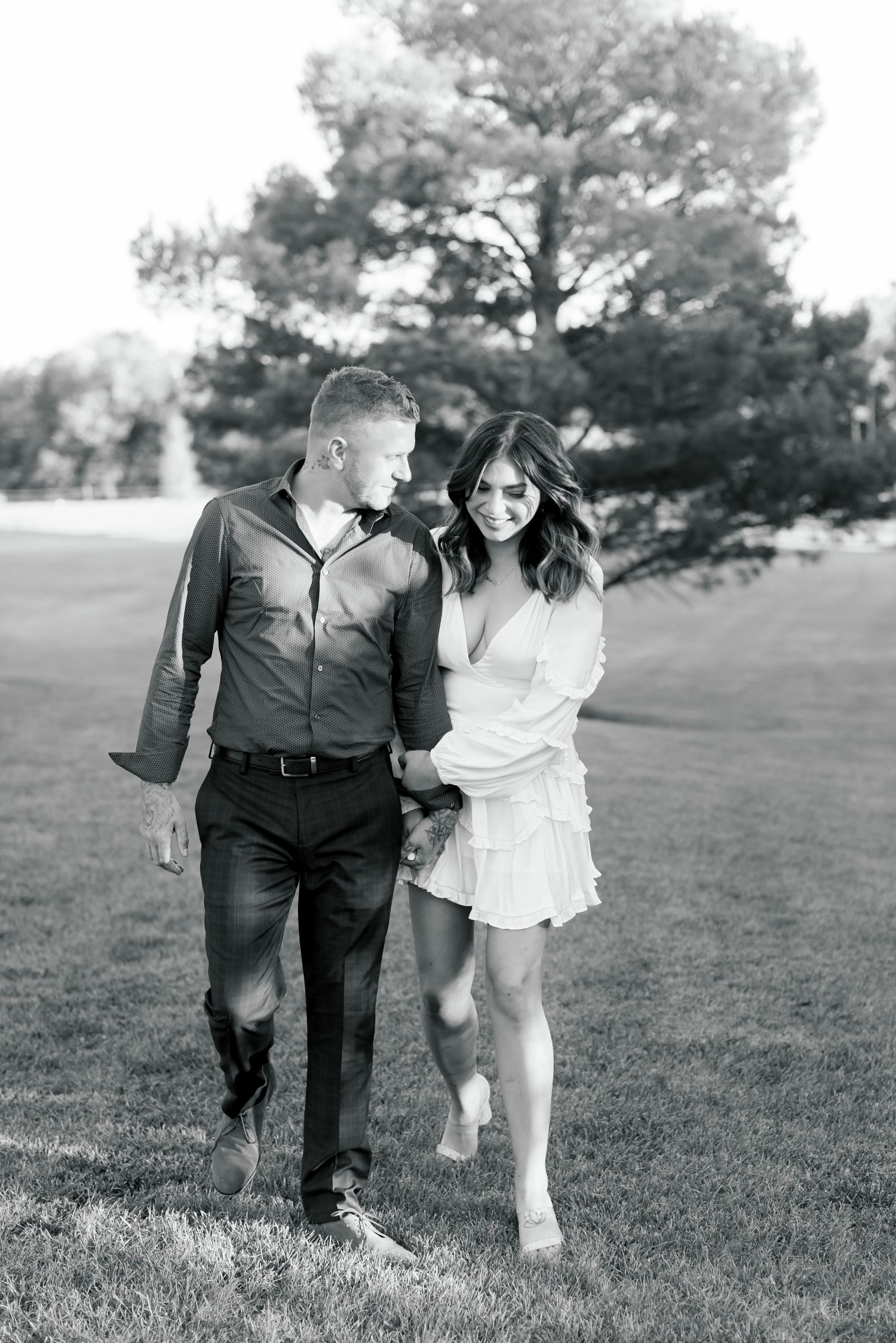 Monochrome photo of couple walking intimately together on grass with pine tree in background.