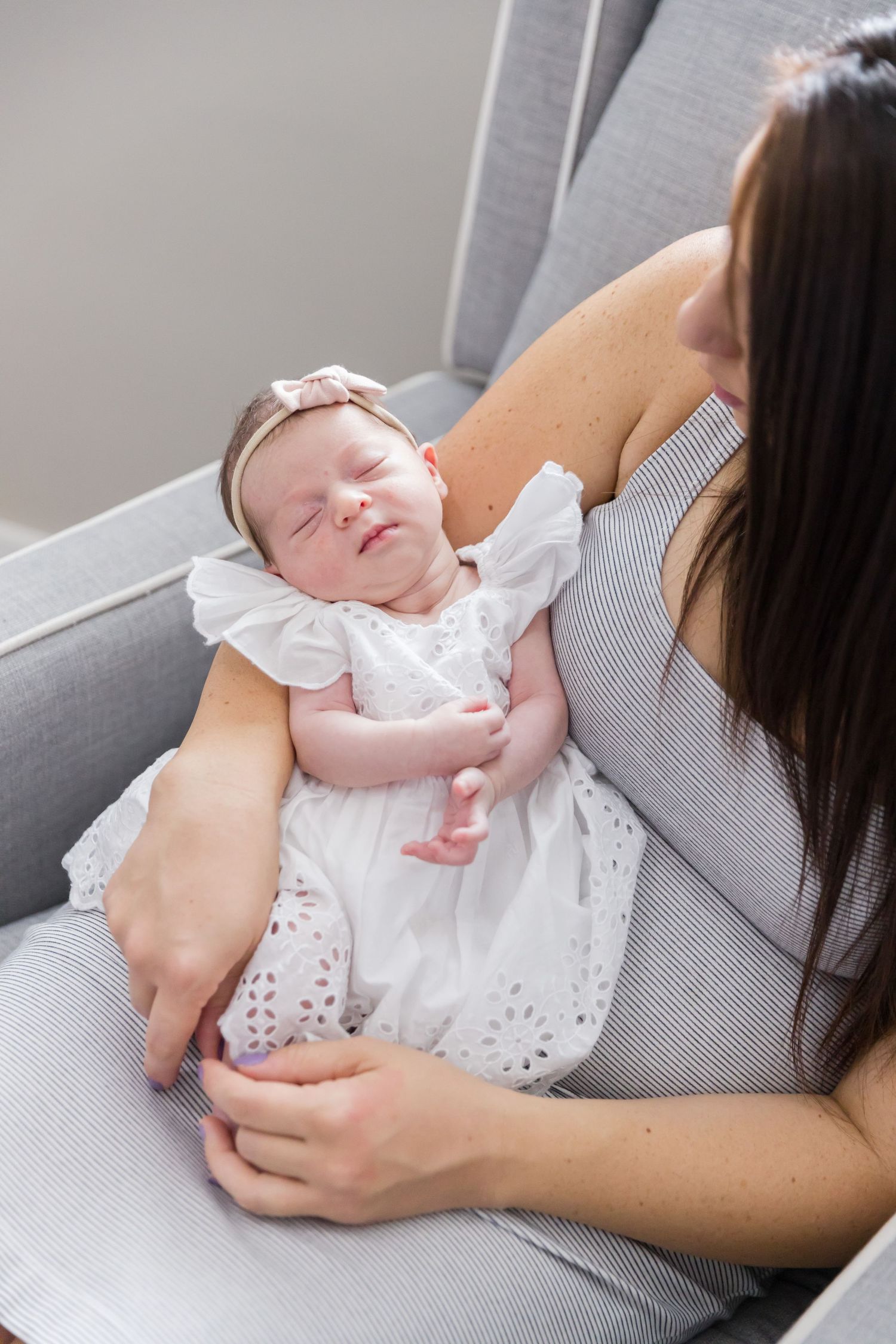 Sleeping newborn in white dress rests peacefully in caregiver's arms on grey chair.