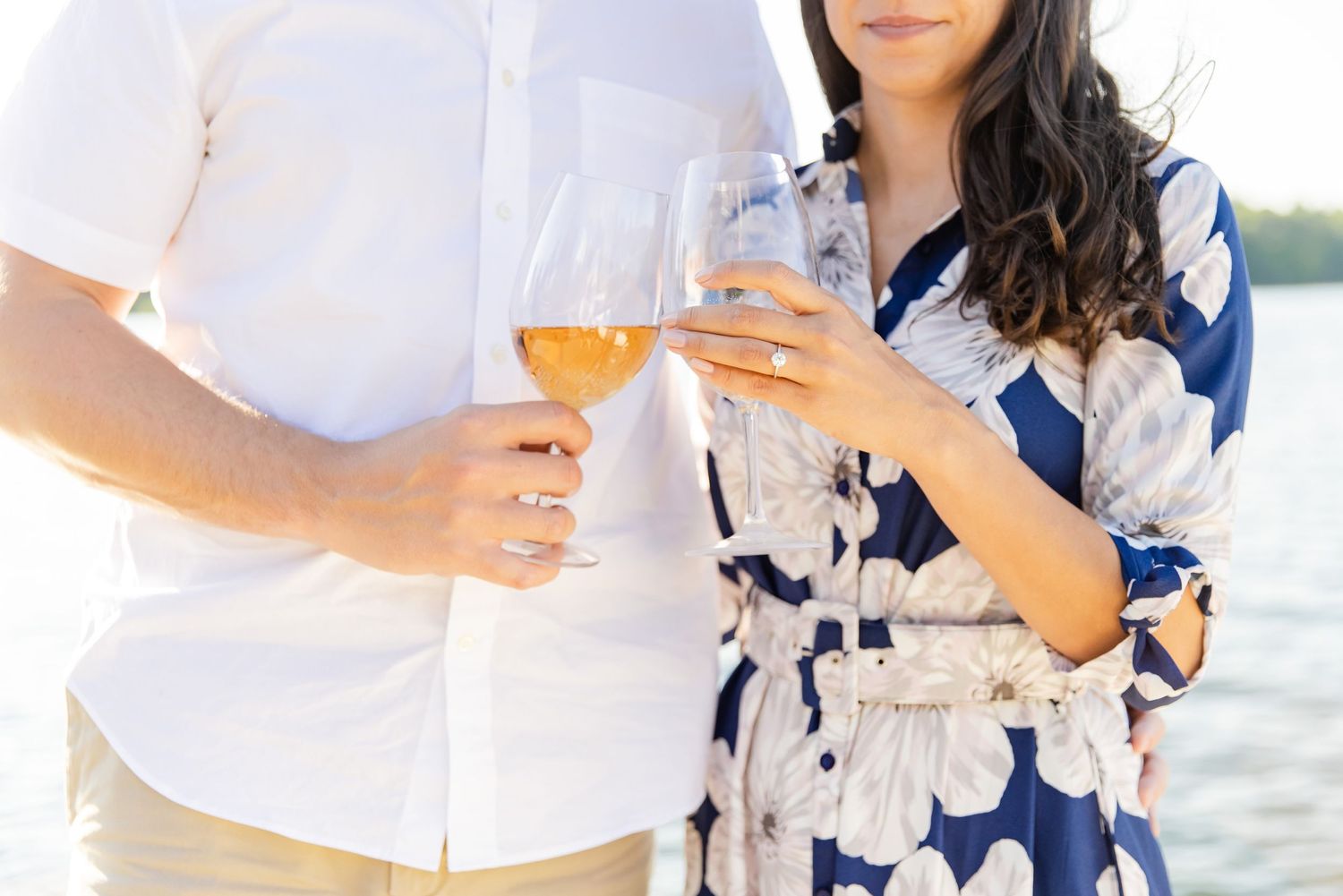 Couple toasting with wine glasses at sunset on a dock wearing coordinated white and blue floral outfits.