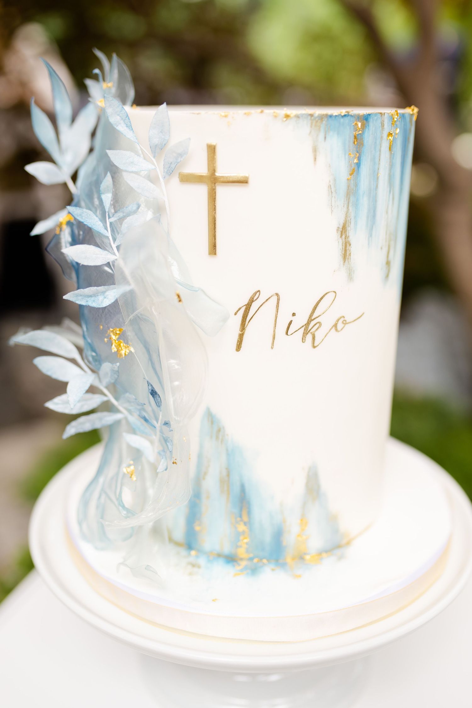 Elegant white baptism cake with gold cross, name 'Niko', and blue watercolor design with white floral details.