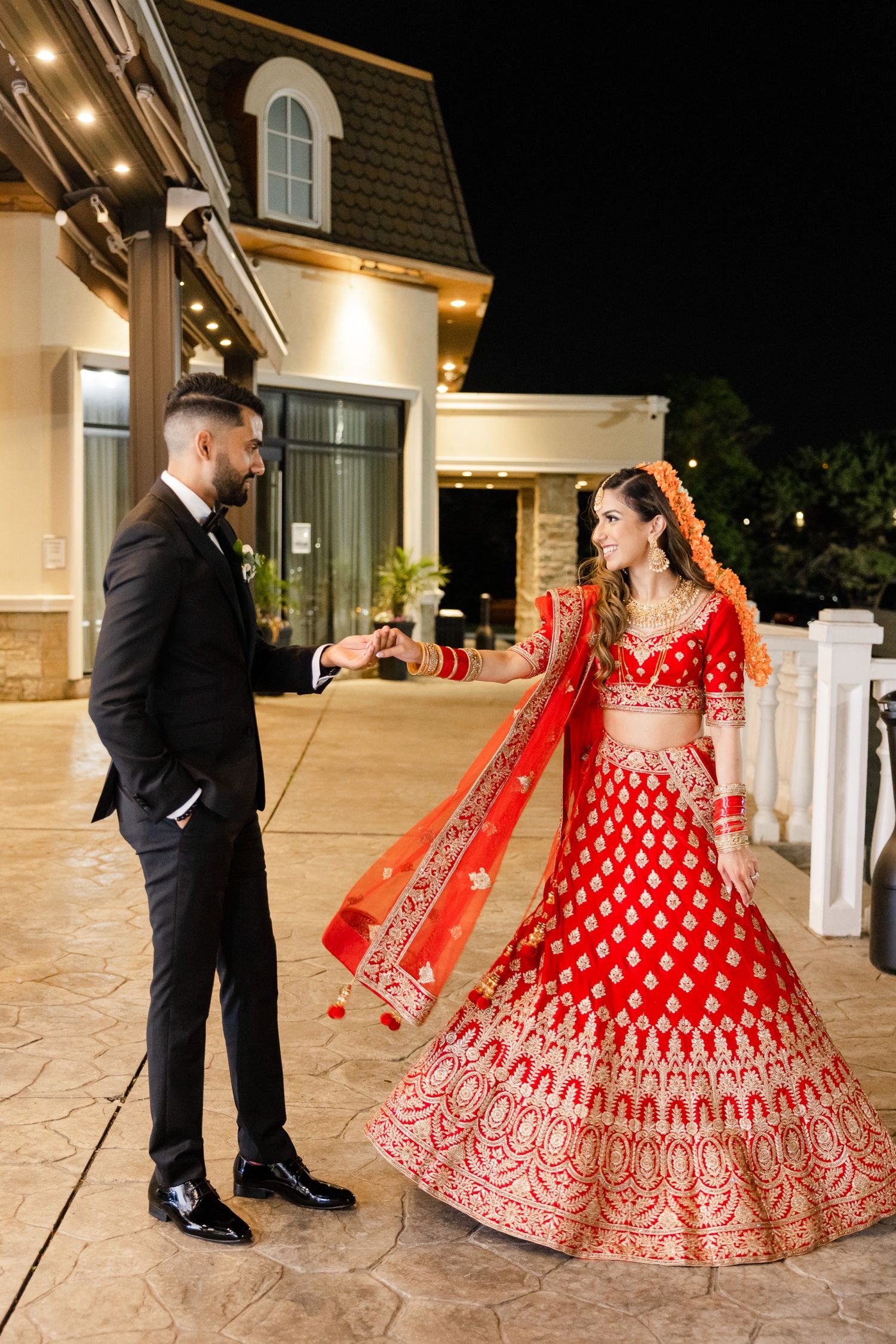 A formal couple poses outdoors, featuring a stunning red and gold embroidered lehenga and black tuxedo.