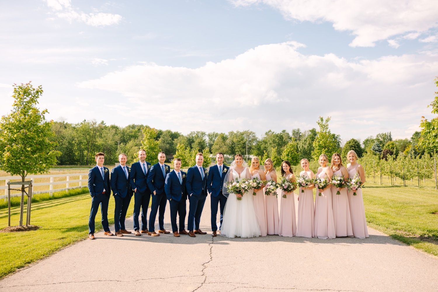 Full wedding party in navy suits and pink dresses poses on a concrete path at an outdoor venue.