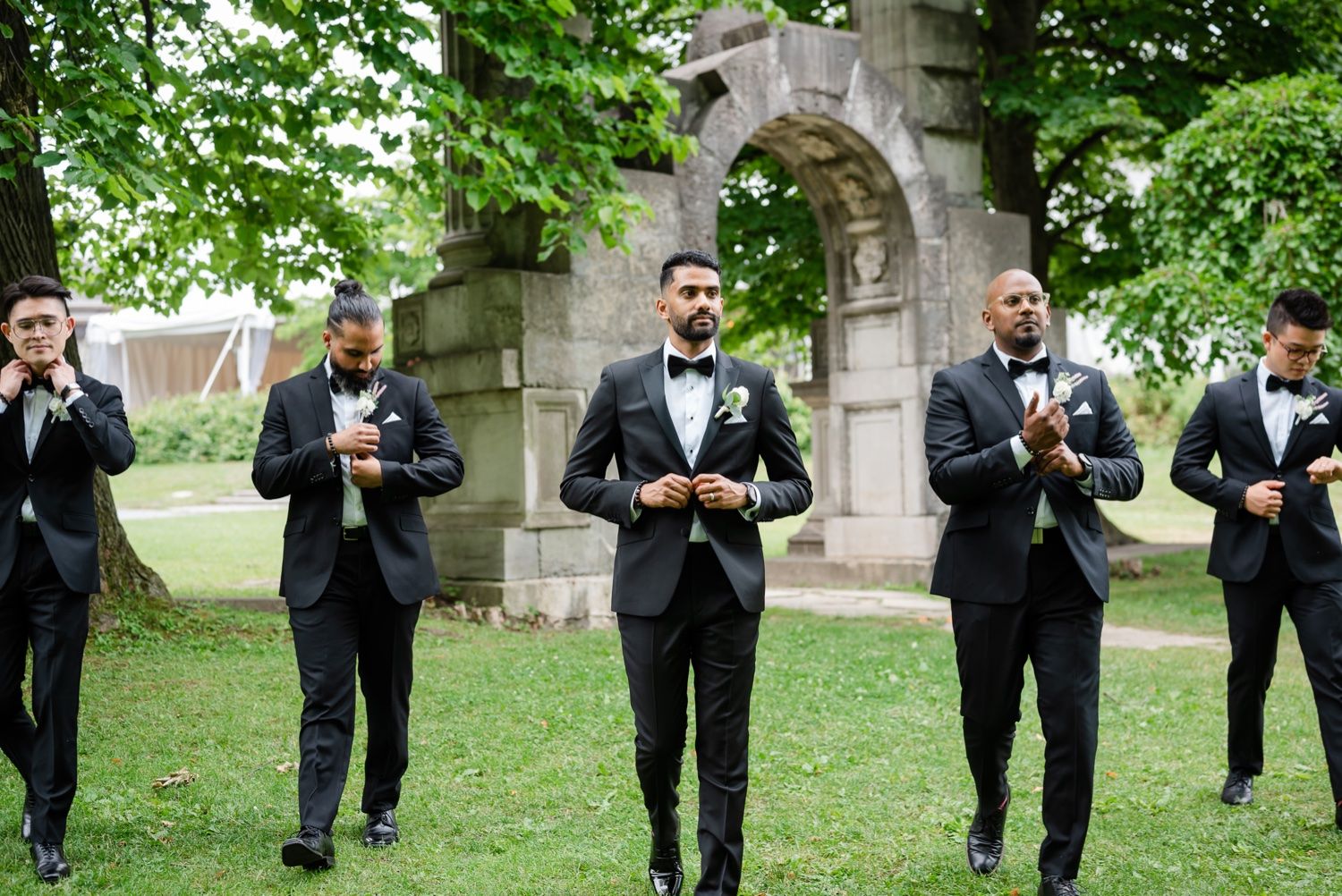 Group of groomsmen checking phones while standing under stone arch in garden.