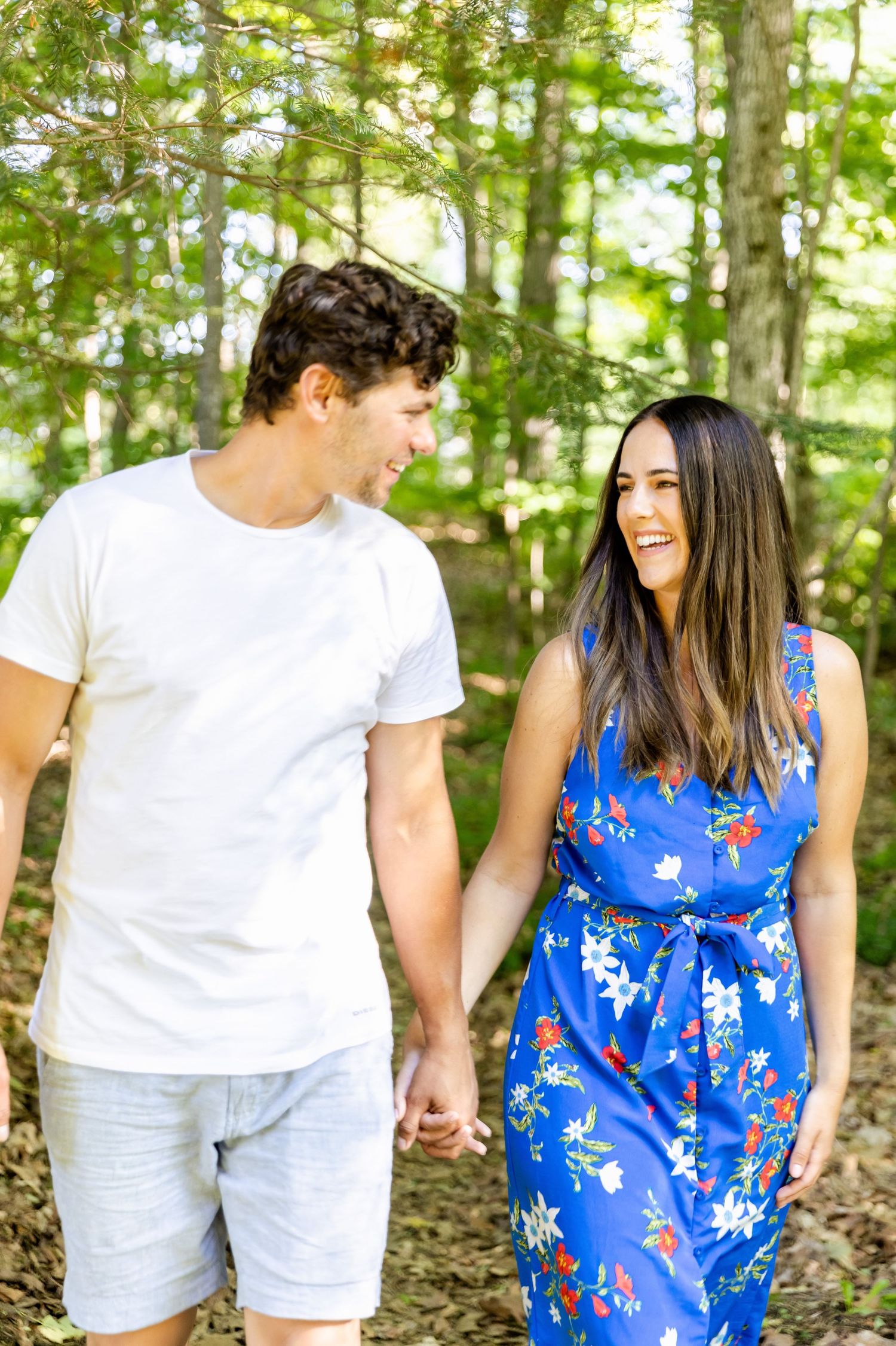 A couple walks hand in hand through a wooded area while sharing a laugh together.
