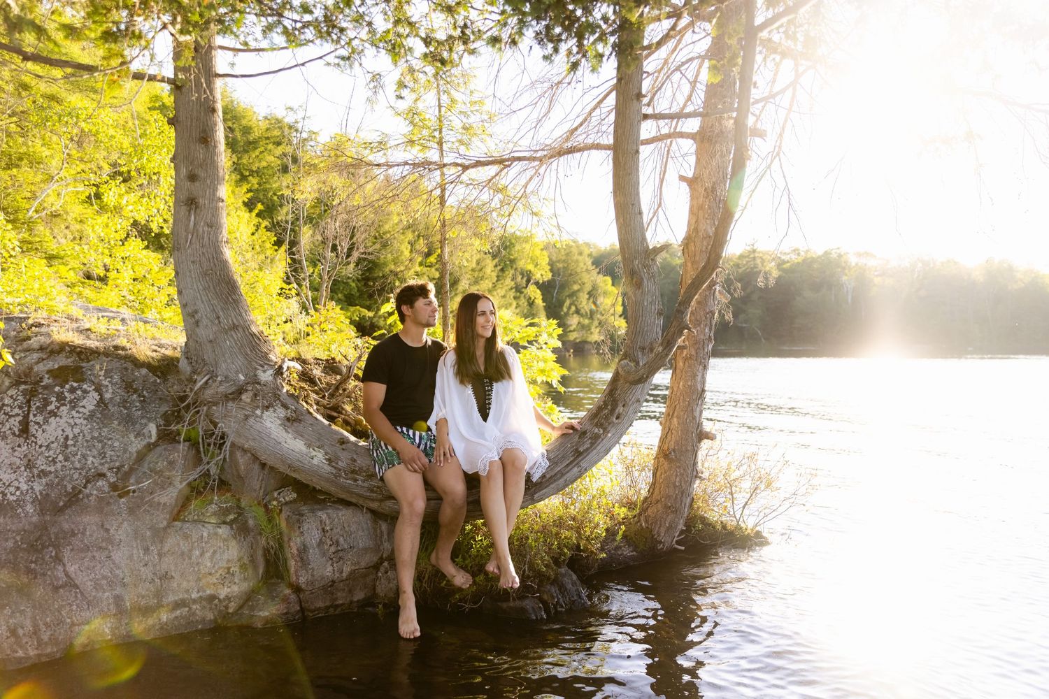 Two people enjoying a peaceful moment on a tree branch overhanging a tranquil lake at sunset.