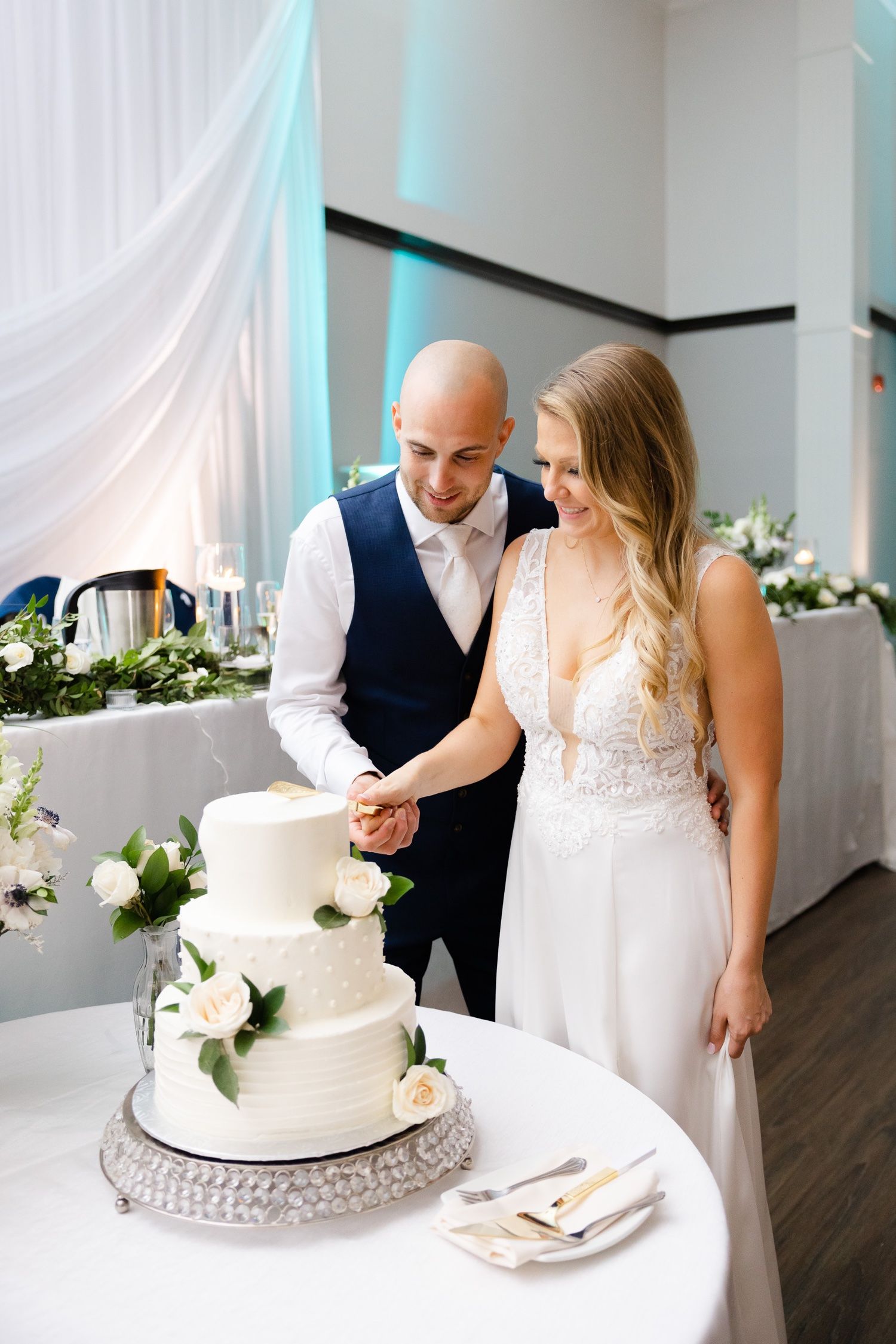A couple cuts their elegant white wedding cake decorated with fresh flowers at their reception.