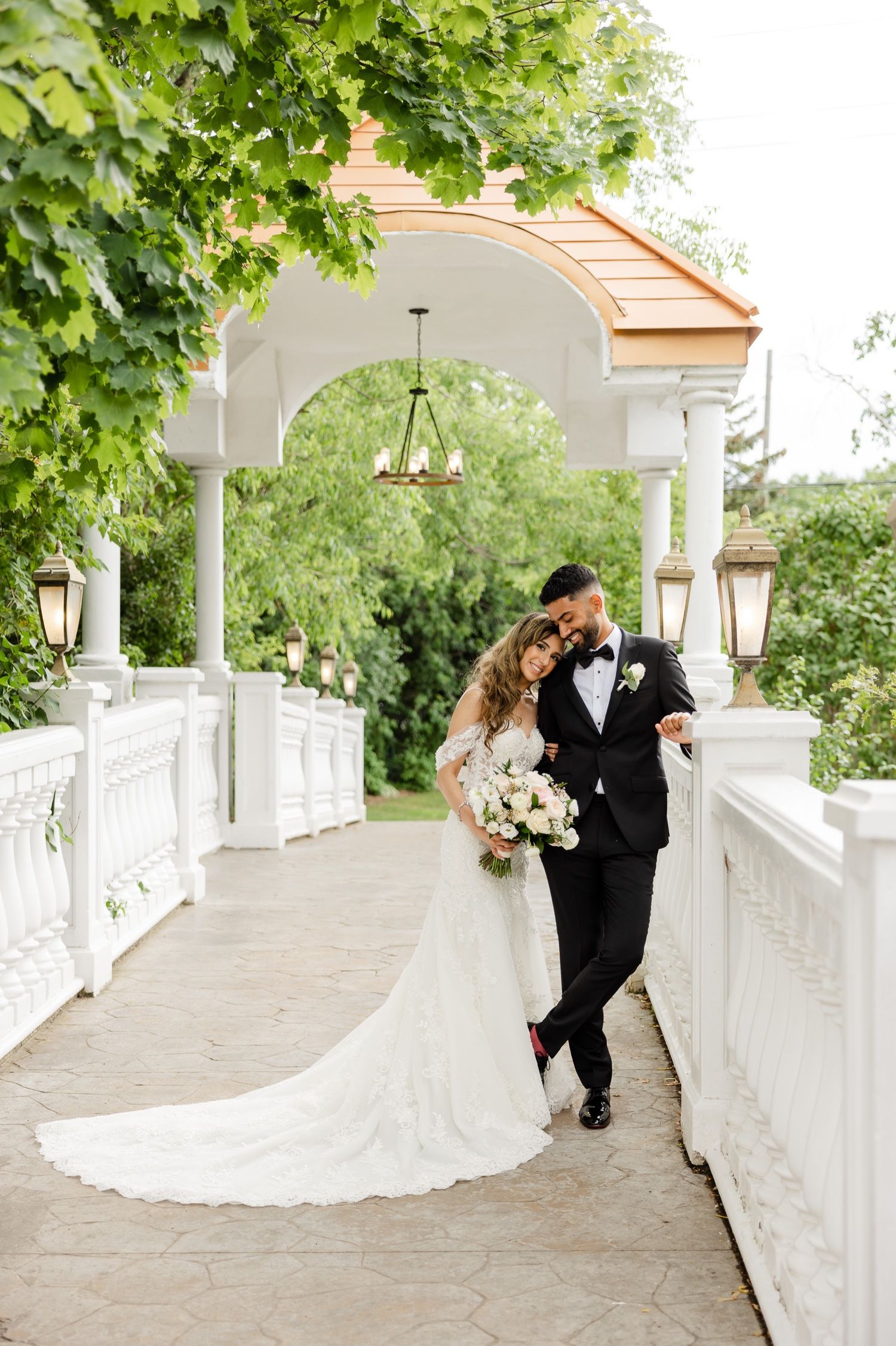 Elegant wedding couple sharing an intimate moment under a white arched walkway with hanging lanterns and lush greenery.