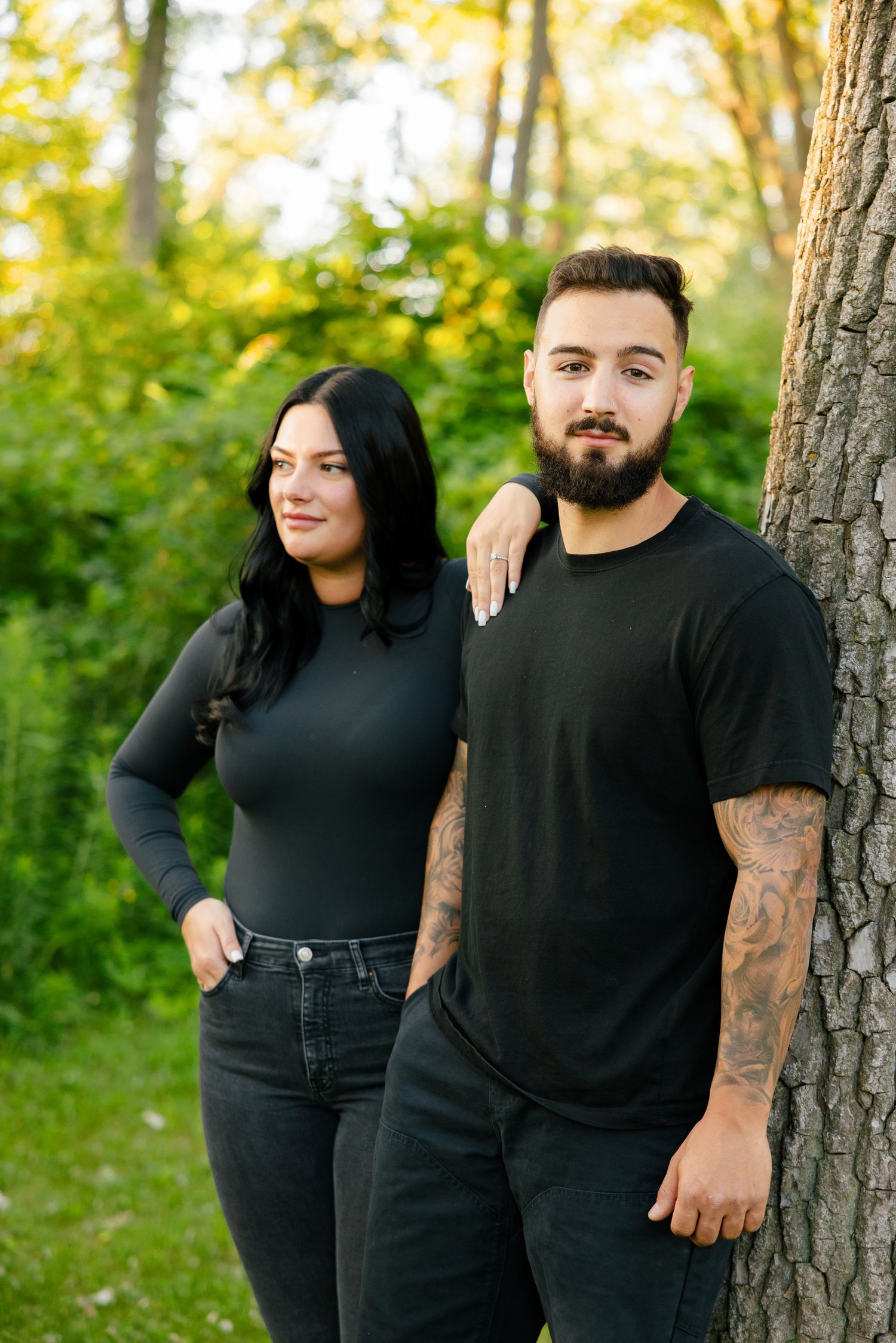 Couple in dark clothing posing against tree trunk with tattoos visible in outdoor forest setting.