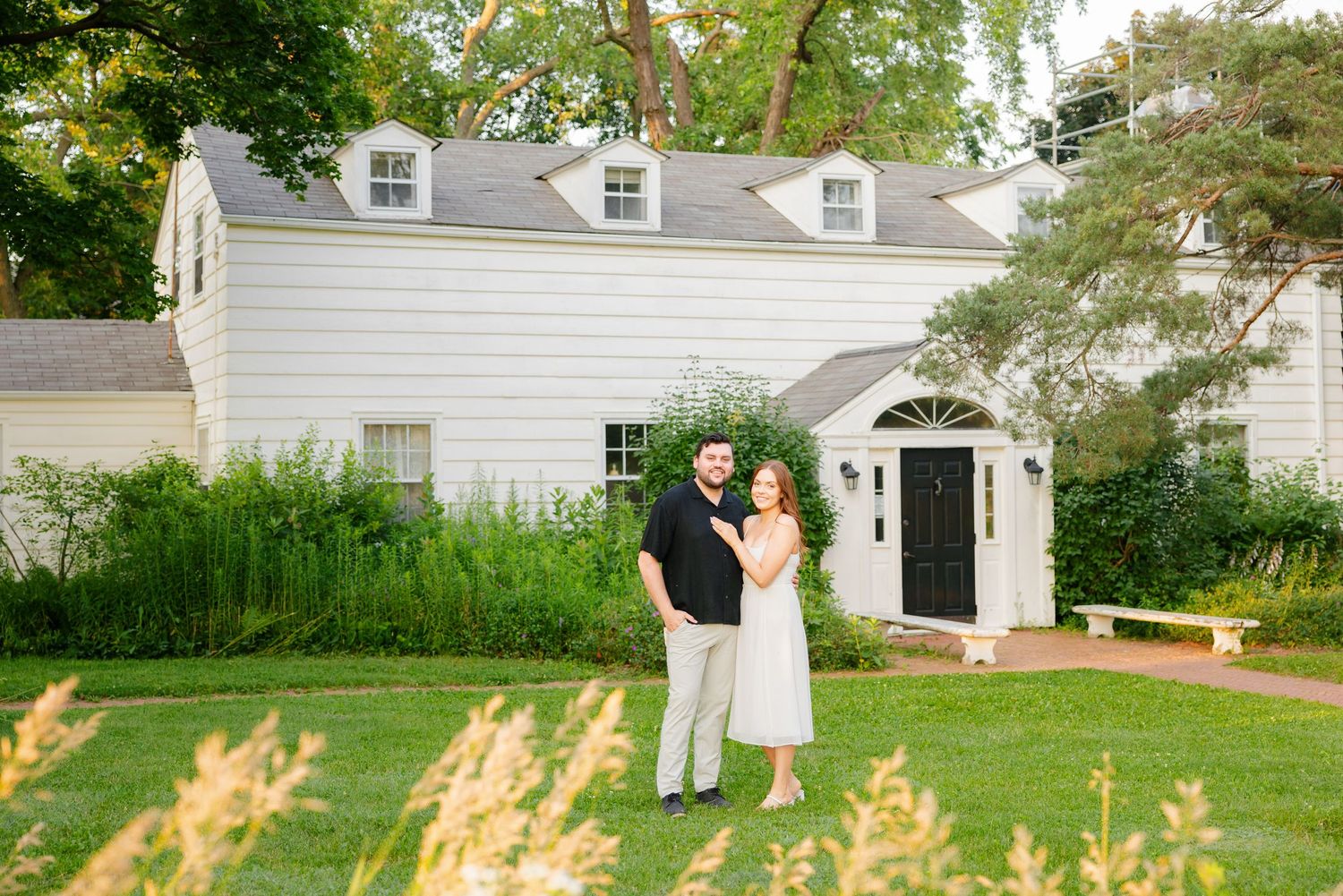 Couple standing together on green lawn with white house and arched doorway in background.