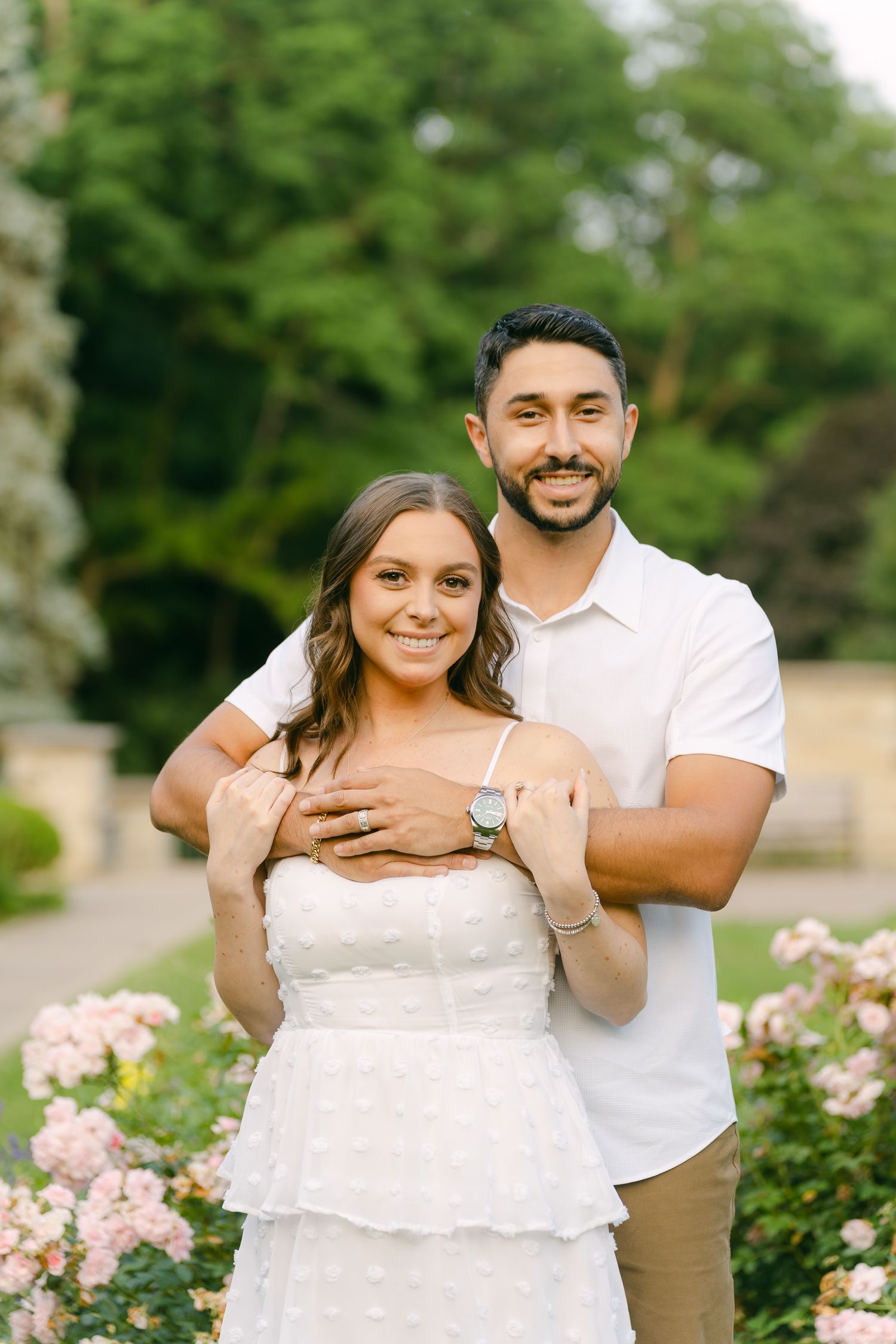 A couple embraces in front of pink rose bushes during their engagement photo session.