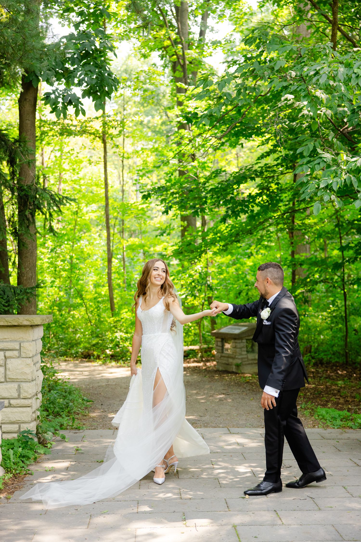 Newlyweds dancing together on stone pathway amidst vibrant green forest scenery.