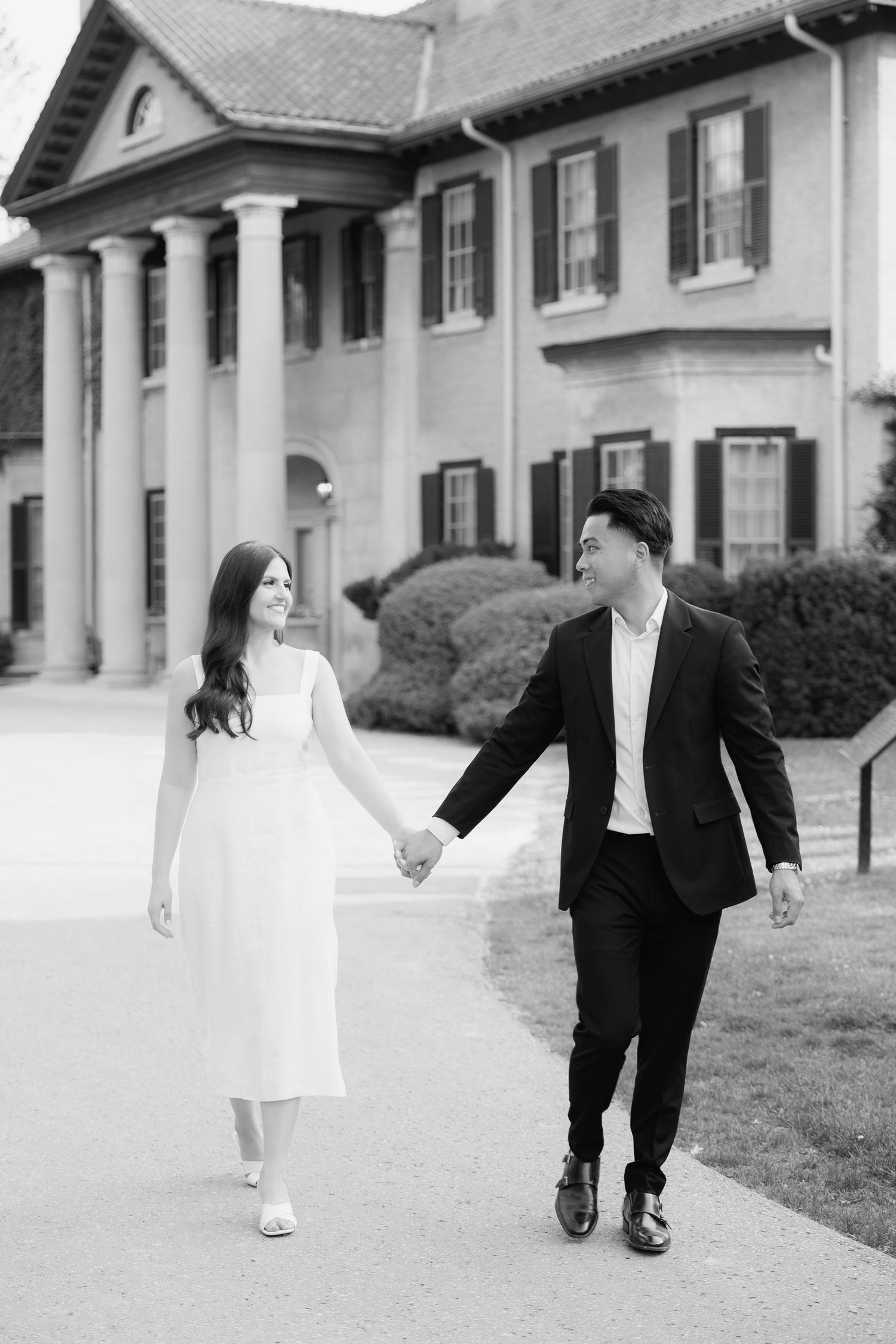Black and white photo of couple walking together by historic mansion entrance.