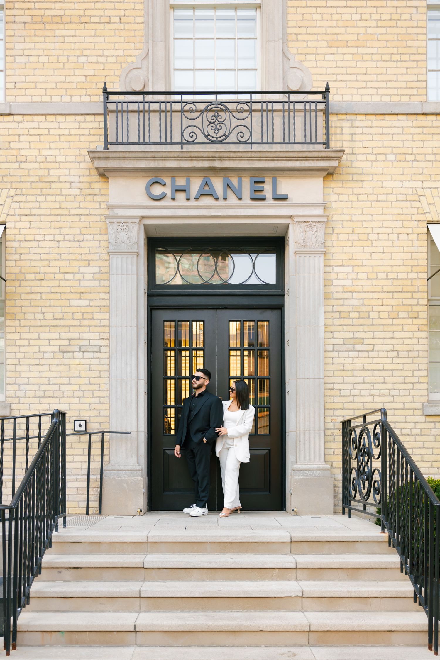 A couple poses at the entrance of a Chanel store with cream brick facade and black doors.