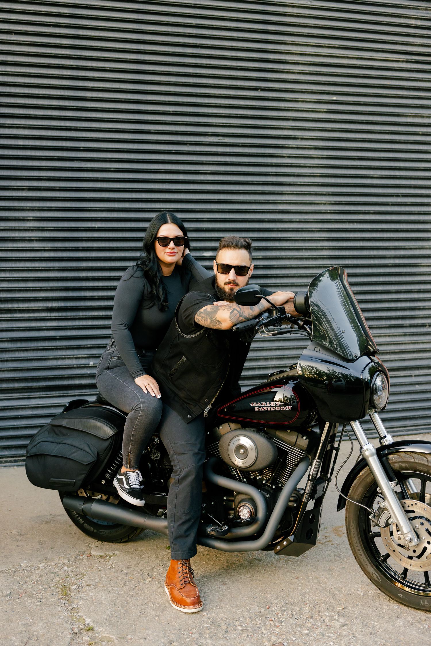 A couple in matching dark outfits pose together on their Harley Davidson motorcycle.