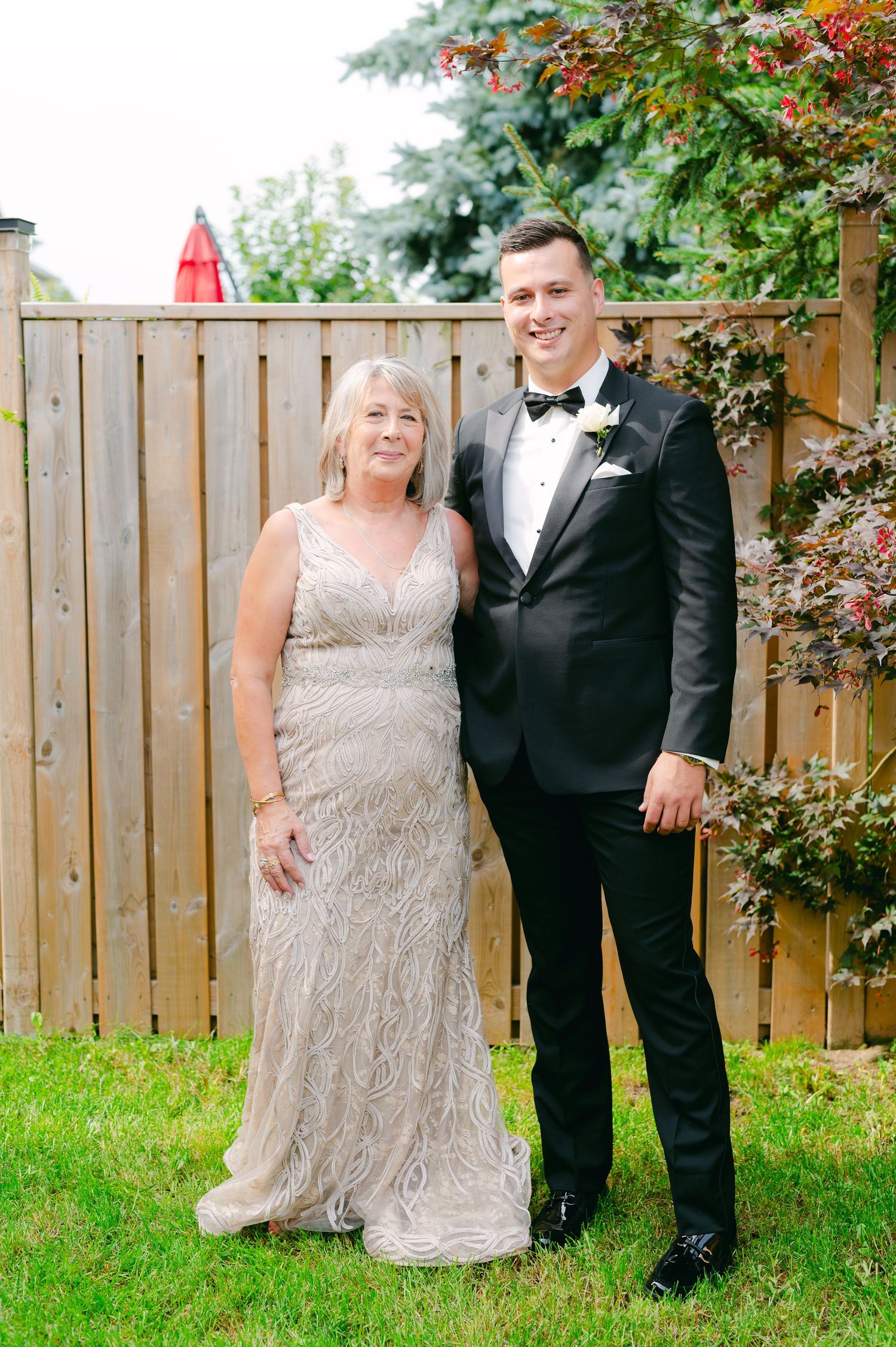 A couple poses together against a wooden fence wearing formal evening attire.