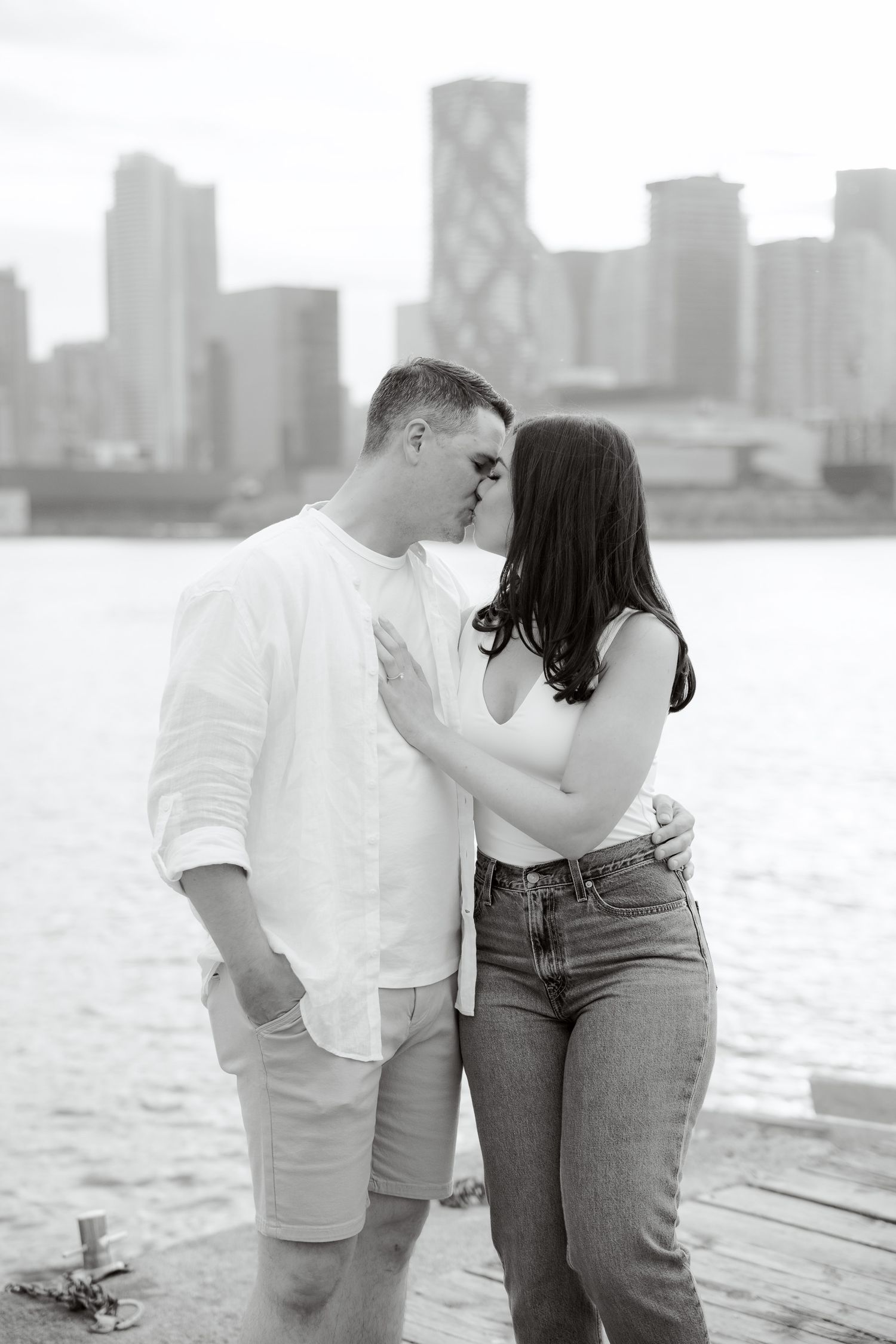 Black and white photo of couple in casual attire sharing an intimate moment on pier with city backdrop.
