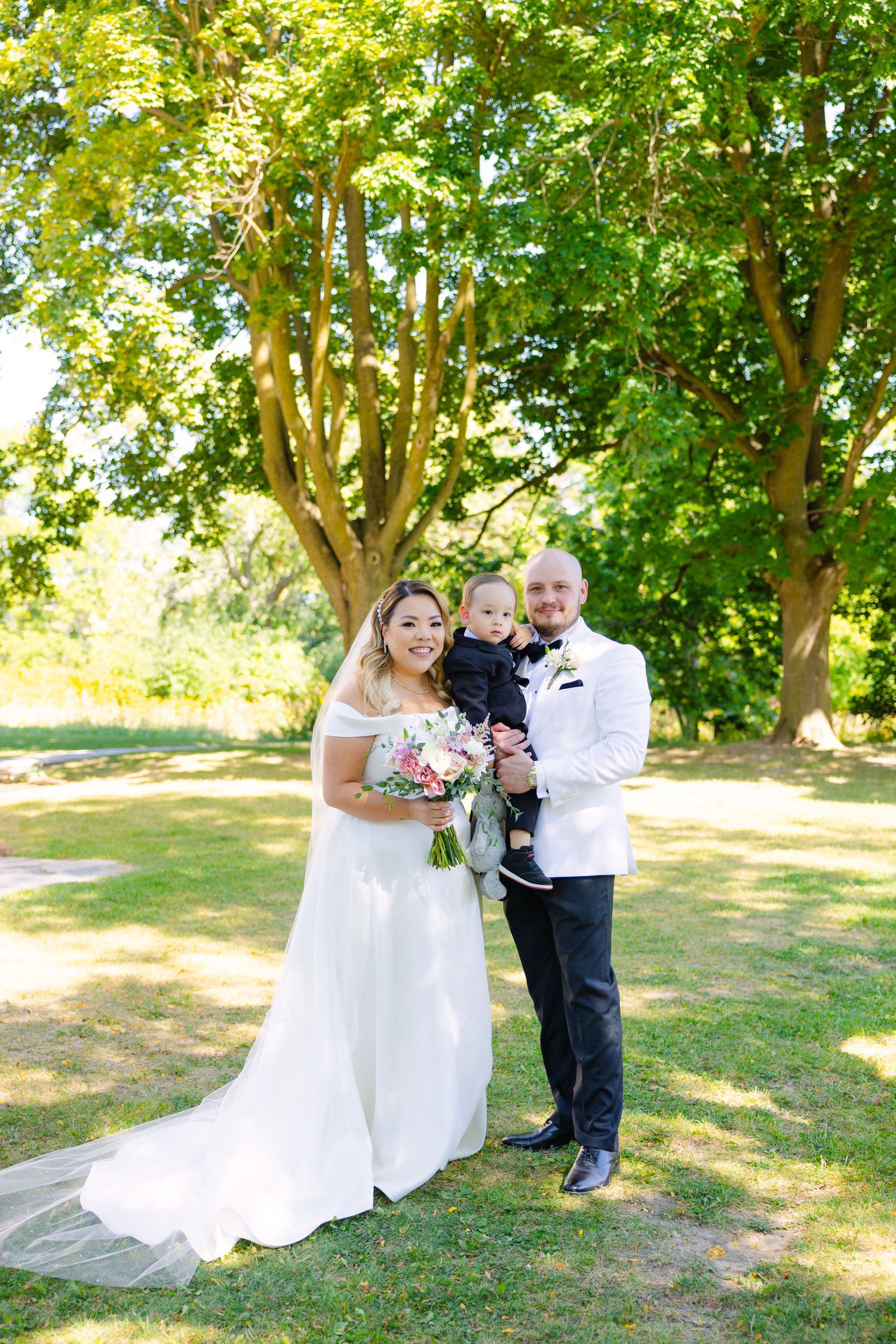 Sunny outdoor wedding photo with couple and young child amongst lush greenery.
