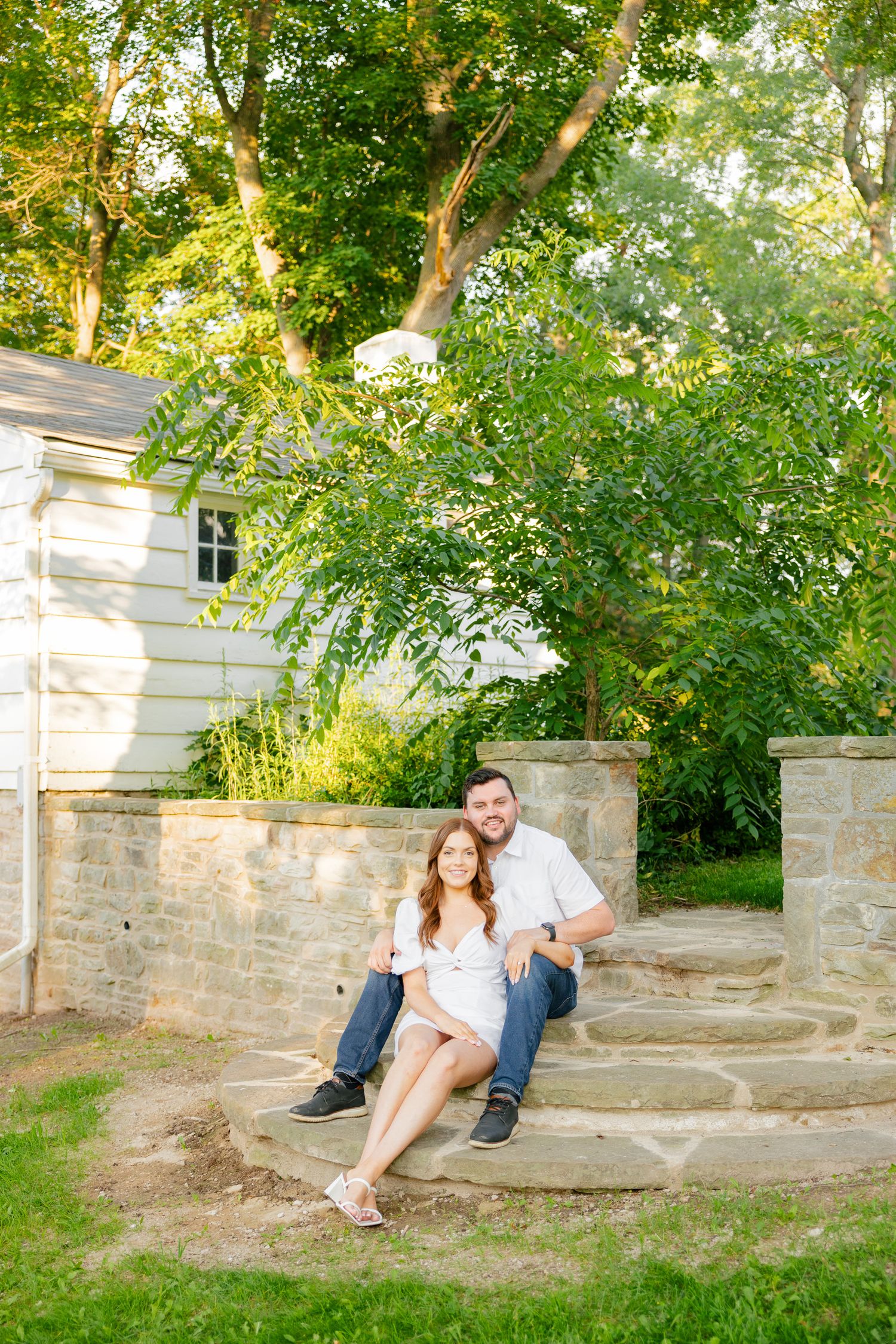 A couple relaxes on stone steps outside a white house surrounded by green foliage.