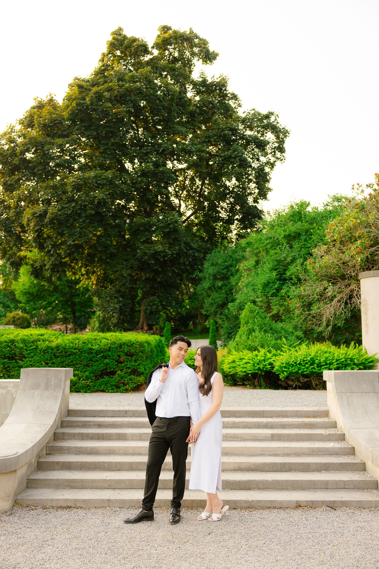 Elegant couple stands together on stone steps in a formal garden surrounded by lush greenery.