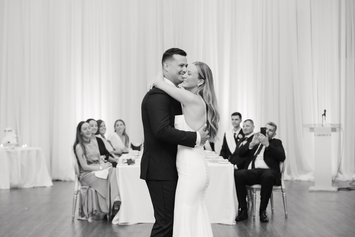 Black and white image of a couple dancing at their wedding reception with seated guests in background.