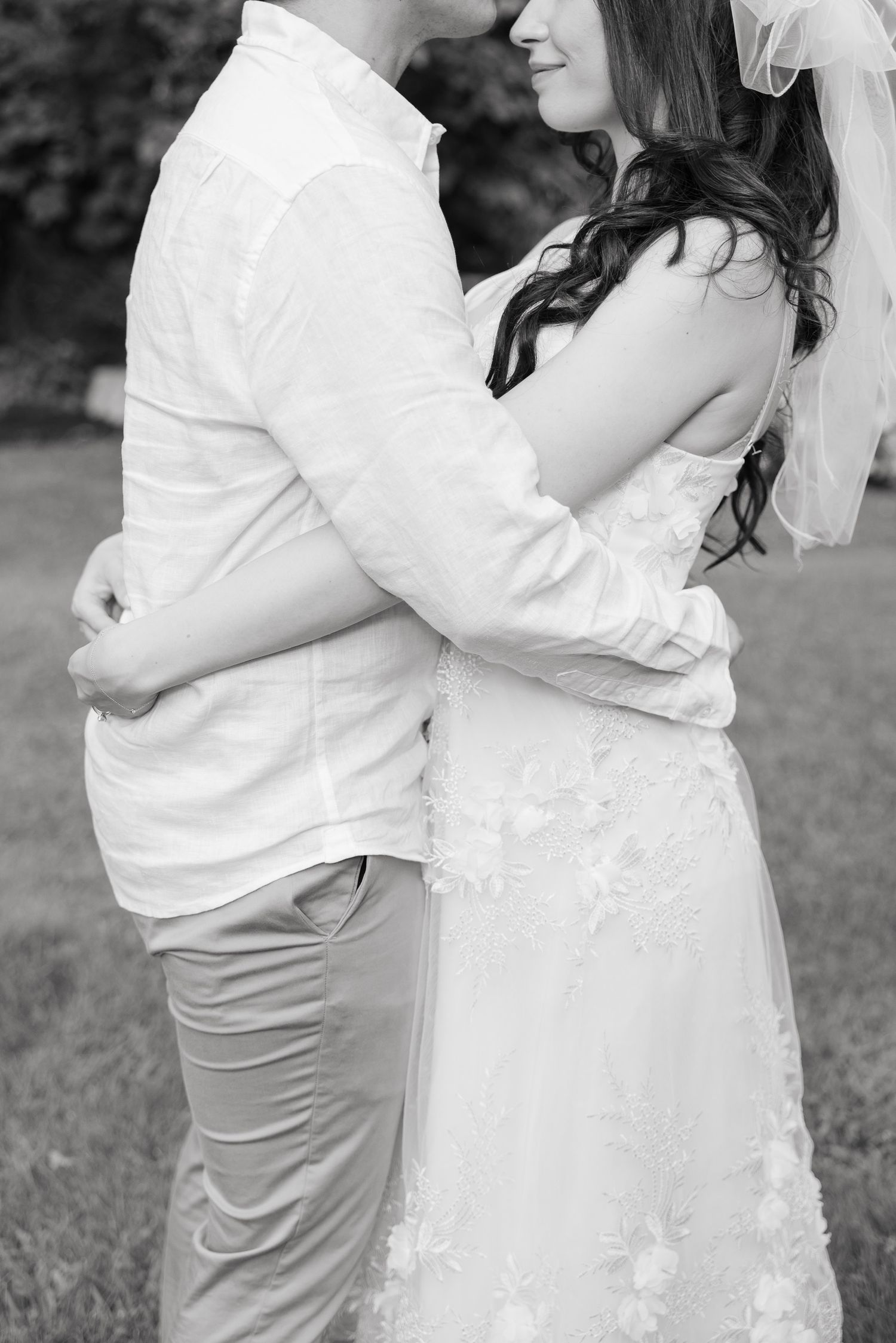 Side view of an embrace between two people in wedding attire standing on grass.