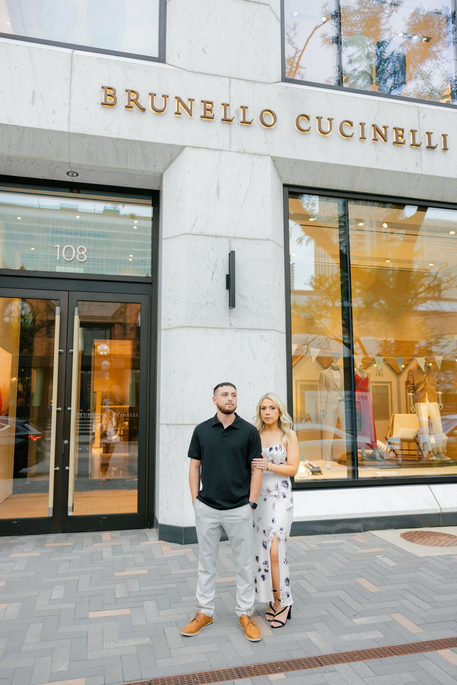 A couple poses in front of the Brunello Cucinelli store entrance wearing coordinated casual attire.