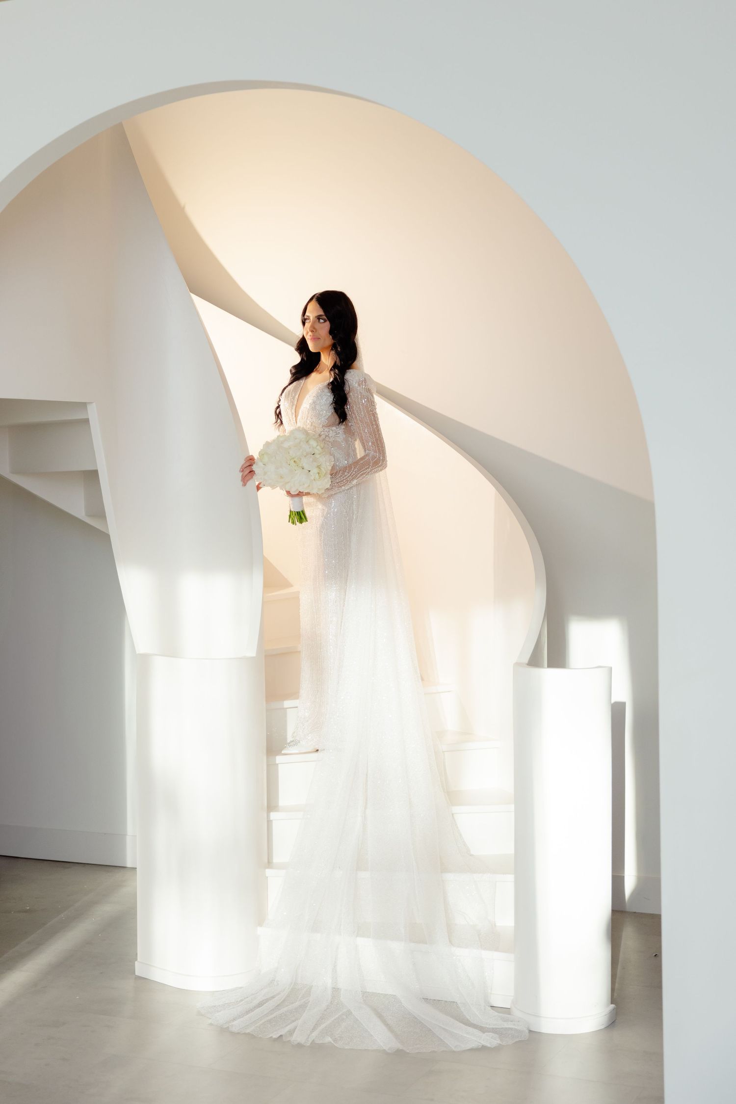 A bride in a fitted lace gown poses elegantly on white stairs beneath a curved archway.