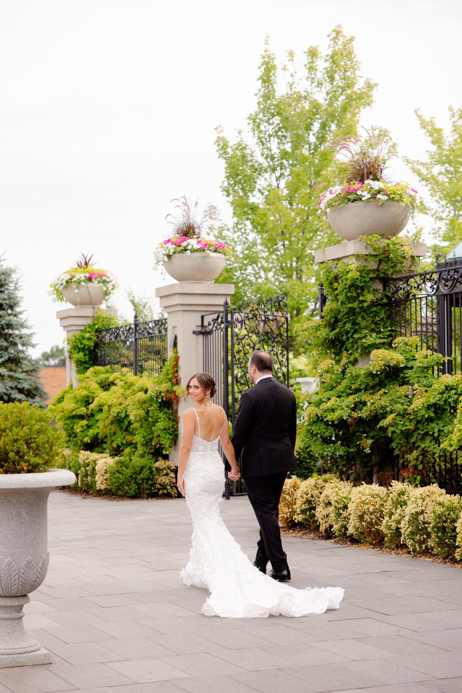 A couple in wedding attire walks along an elegant garden path with decorative stone pillars and flower planters.