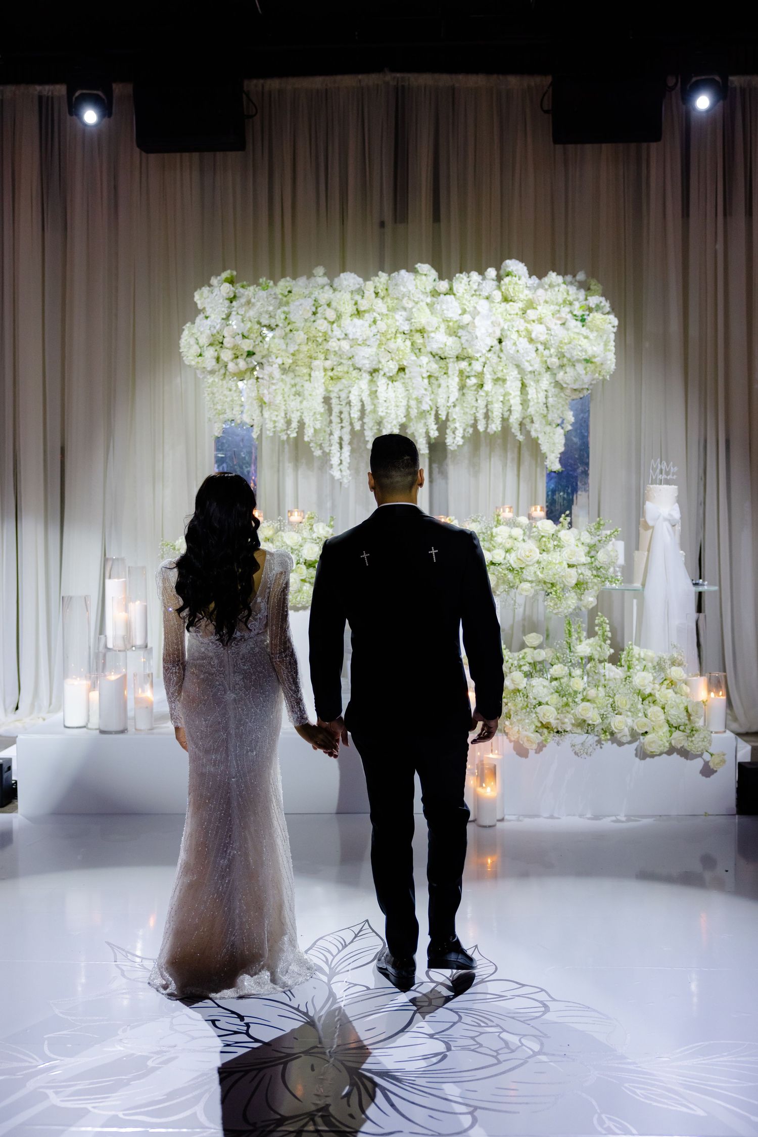 A couple stands silhouetted under an elegant white floral arch at their wedding ceremony.