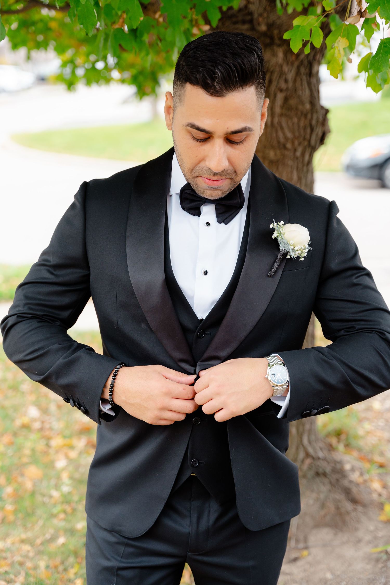 Groom adjusting his tuxedo jacket button while standing under a tree.