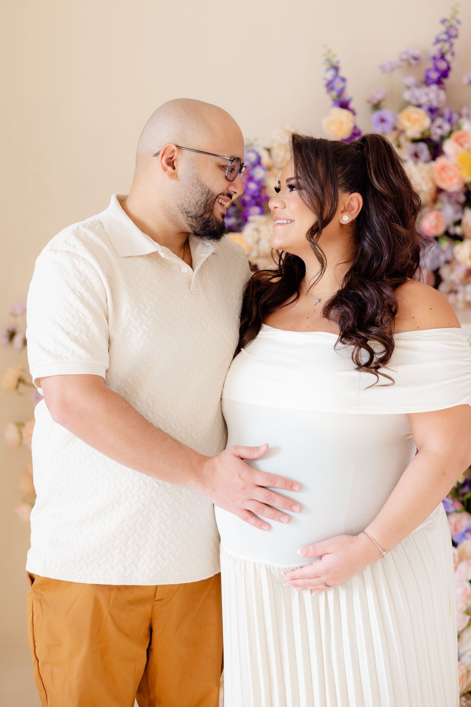 A couple poses for a maternity photo against a floral backdrop wearing white outfits and showing affection.