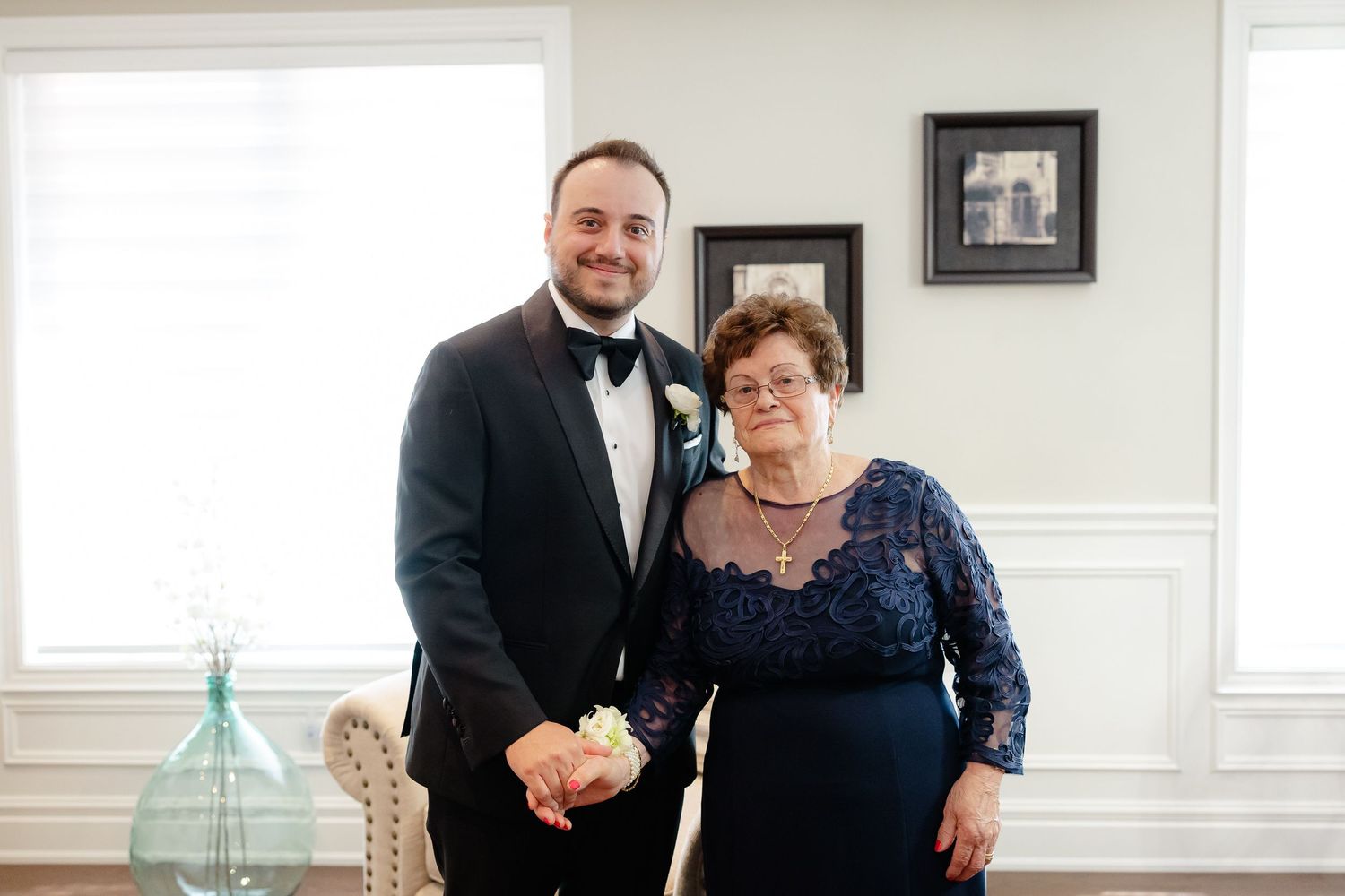Two people pose together in formal attire, with a navy lace dress and black tuxedo, inside an elegant white room.