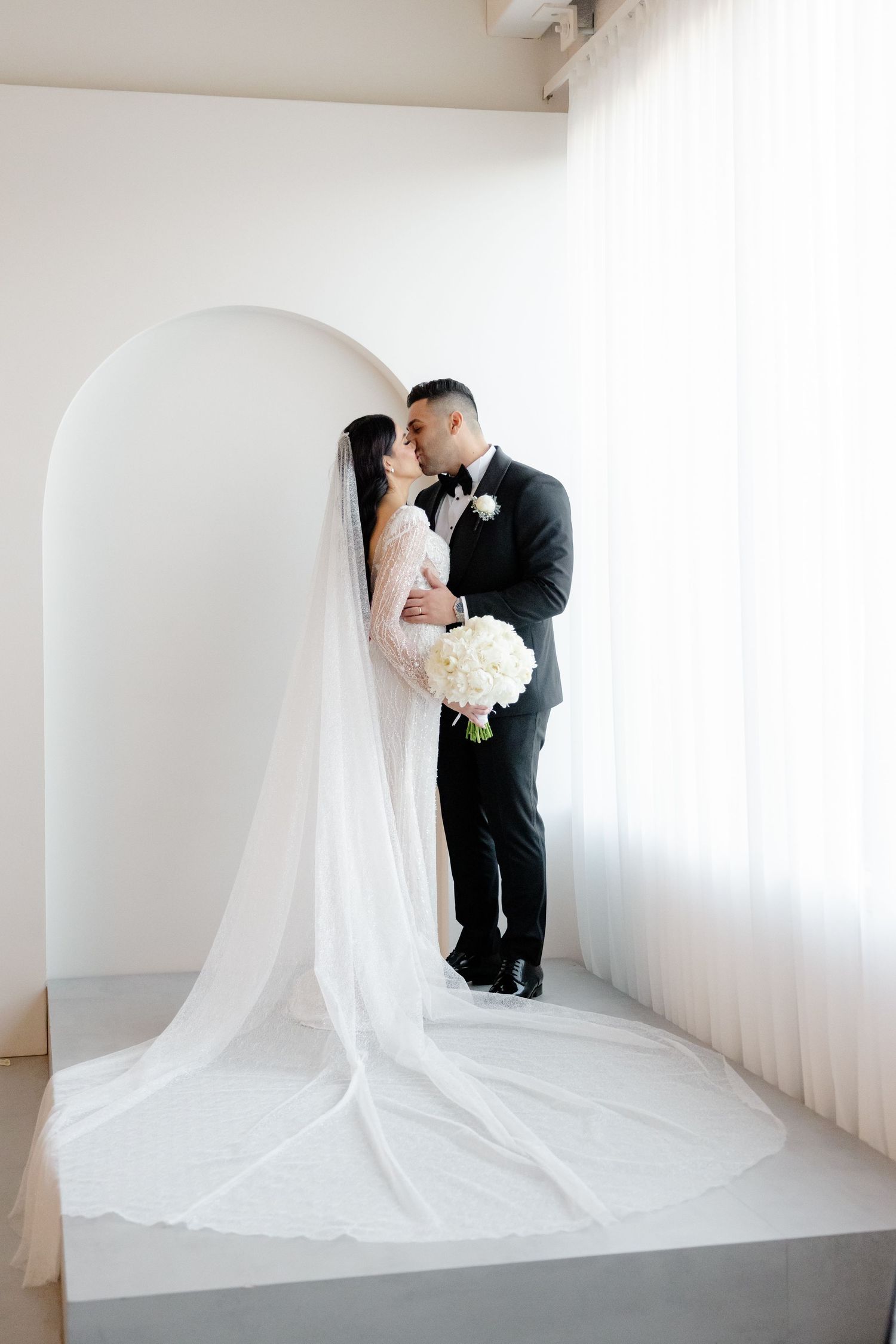 Elegant wedding couple poses against white archway with cathedral veil trailing dramatically.