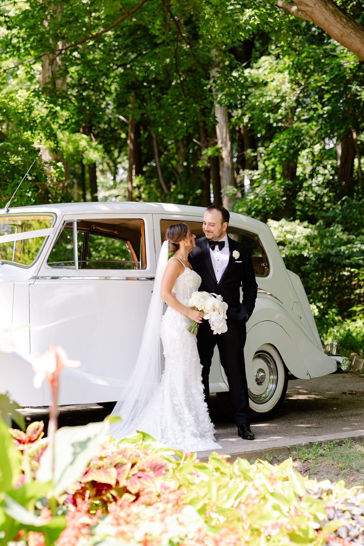 A couple in formalwear stands beside a vintage white car on a tree-lined path during a wedding celebration.