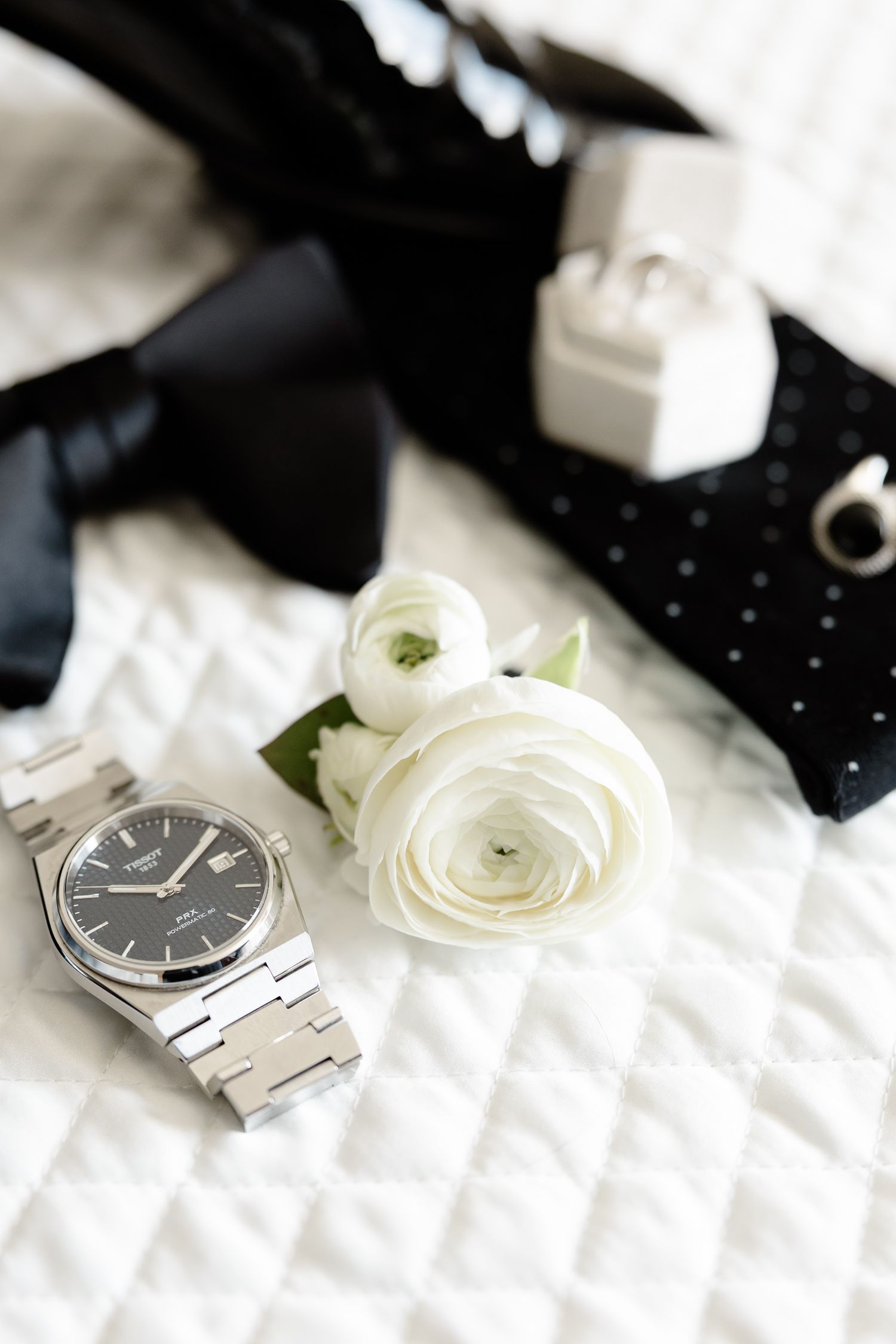 A luxury silver watch with metal band lies beside white ranunculus flowers and a black bow tie on textured fabric.