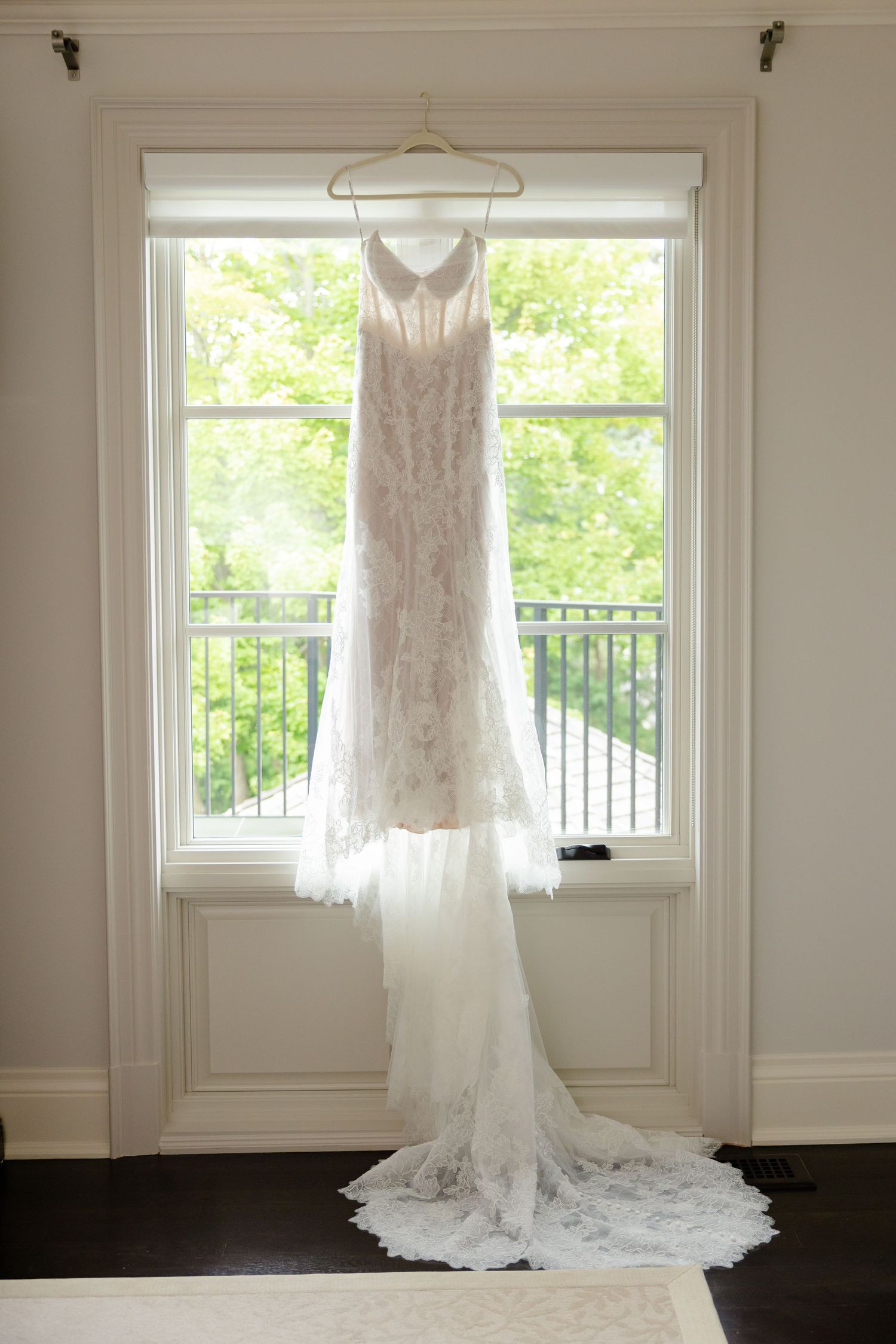 A white lace wedding dress with a long train hangs elegantly in front of a bright window with garden views.