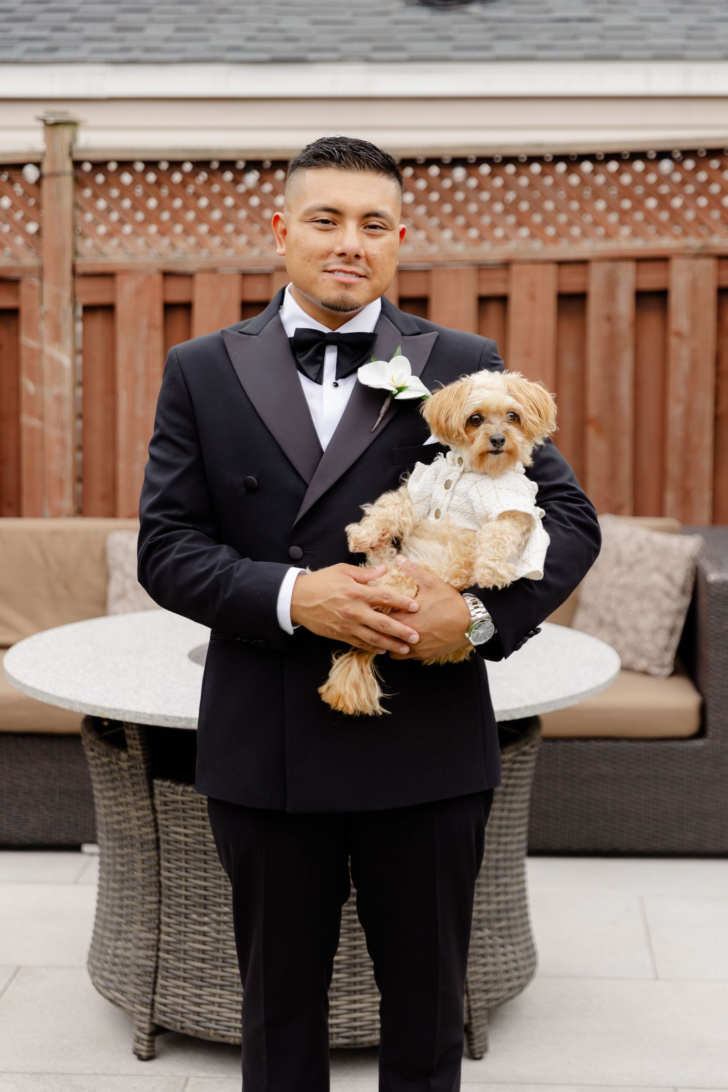 A small dog wearing a white bow tie sits in formal attire with someone in a black suit at an outdoor event.