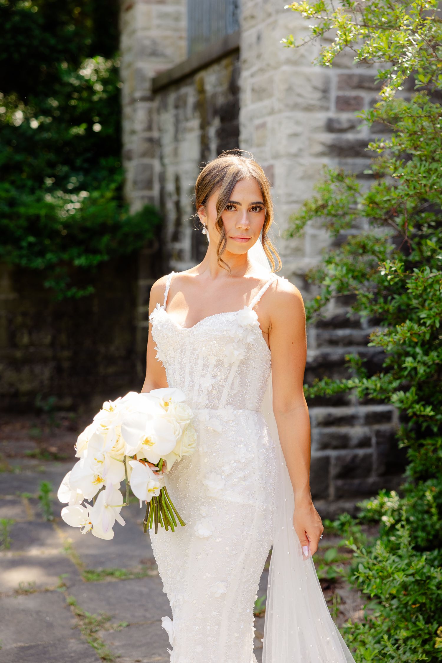Elegant white lace wedding gown with delicate straps photographed against stone wall and greenery.