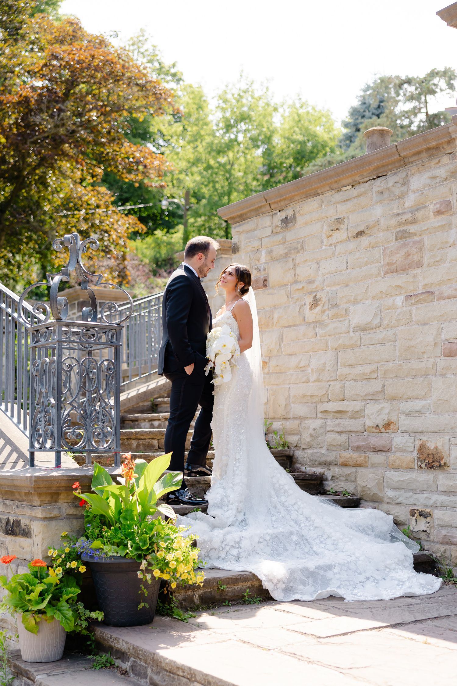 A romantic wedding scene set against a stone wall with autumn foliage and decorative planters along a paved pathway.