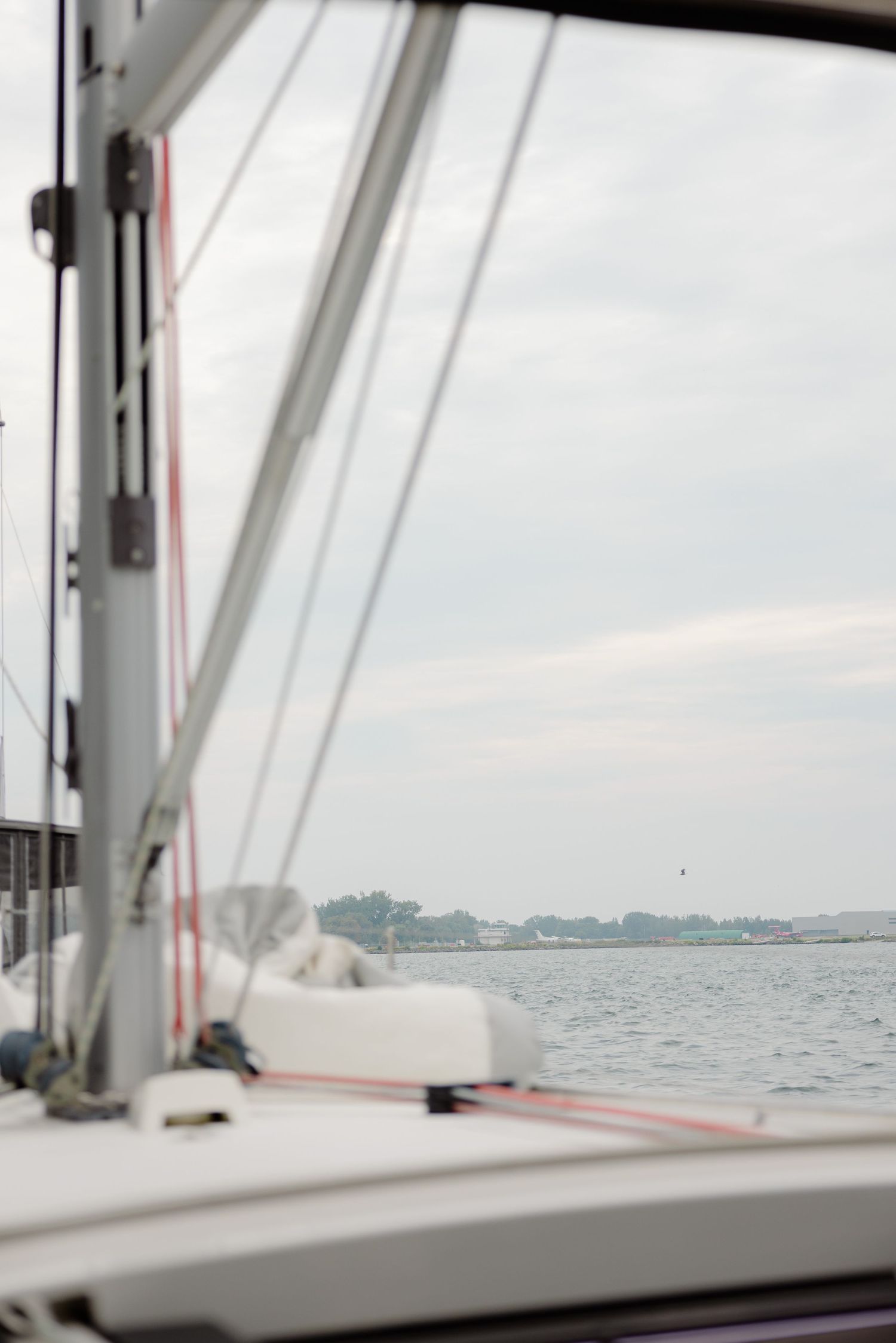 View from a sailboat deck showing rigging lines and calm waters with a distant shoreline on a cloudy day.
