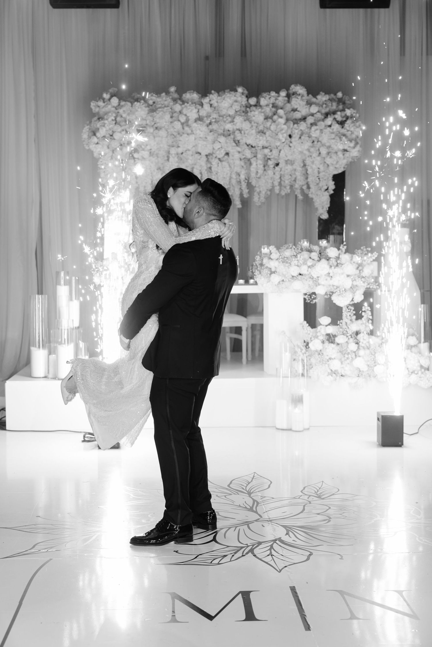 Black and white photo of wedding couple dancing beneath floral arch with sparkler effects.