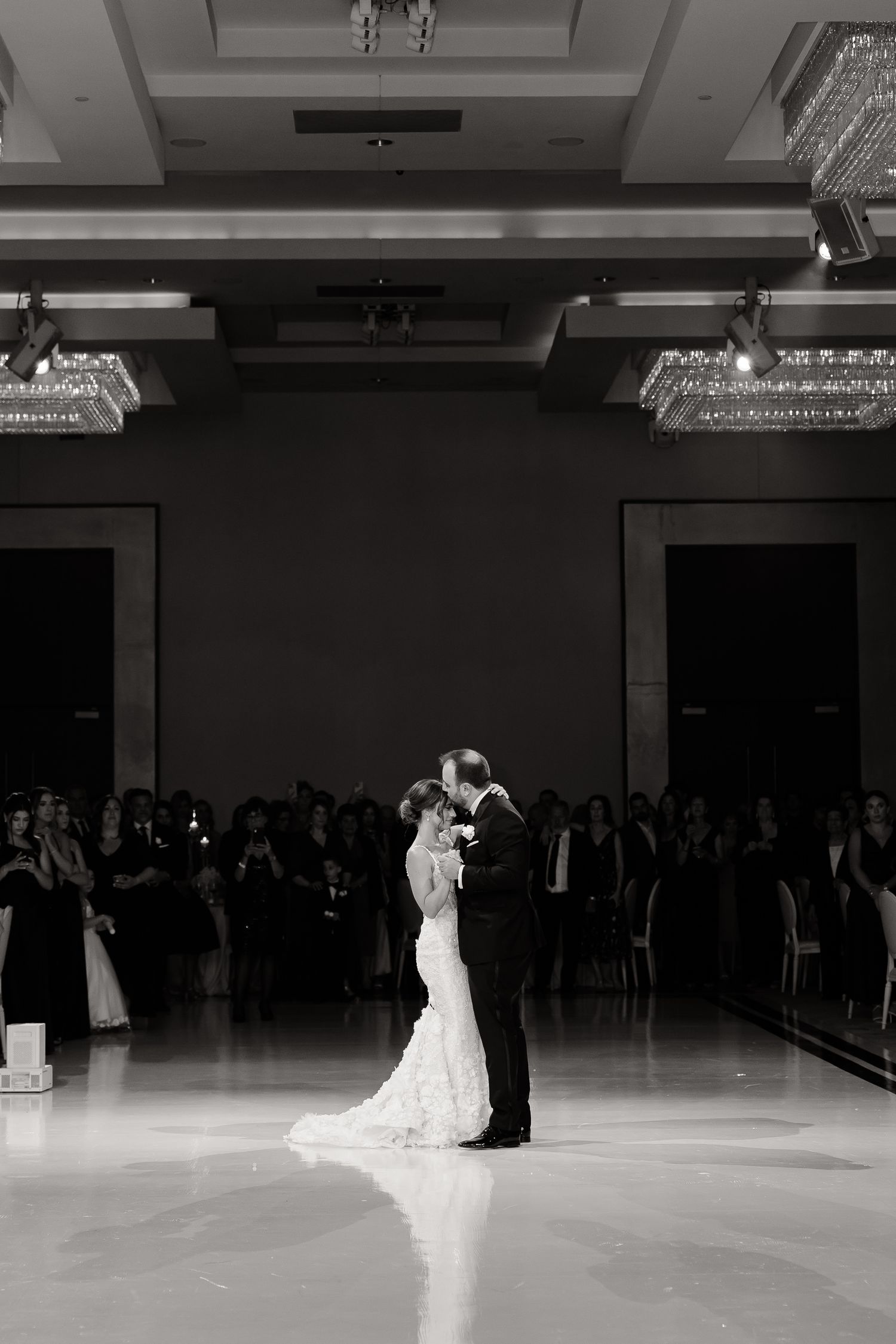 A couple shares their first dance as newlyweds on an elegant ballroom floor under crystal chandeliers.