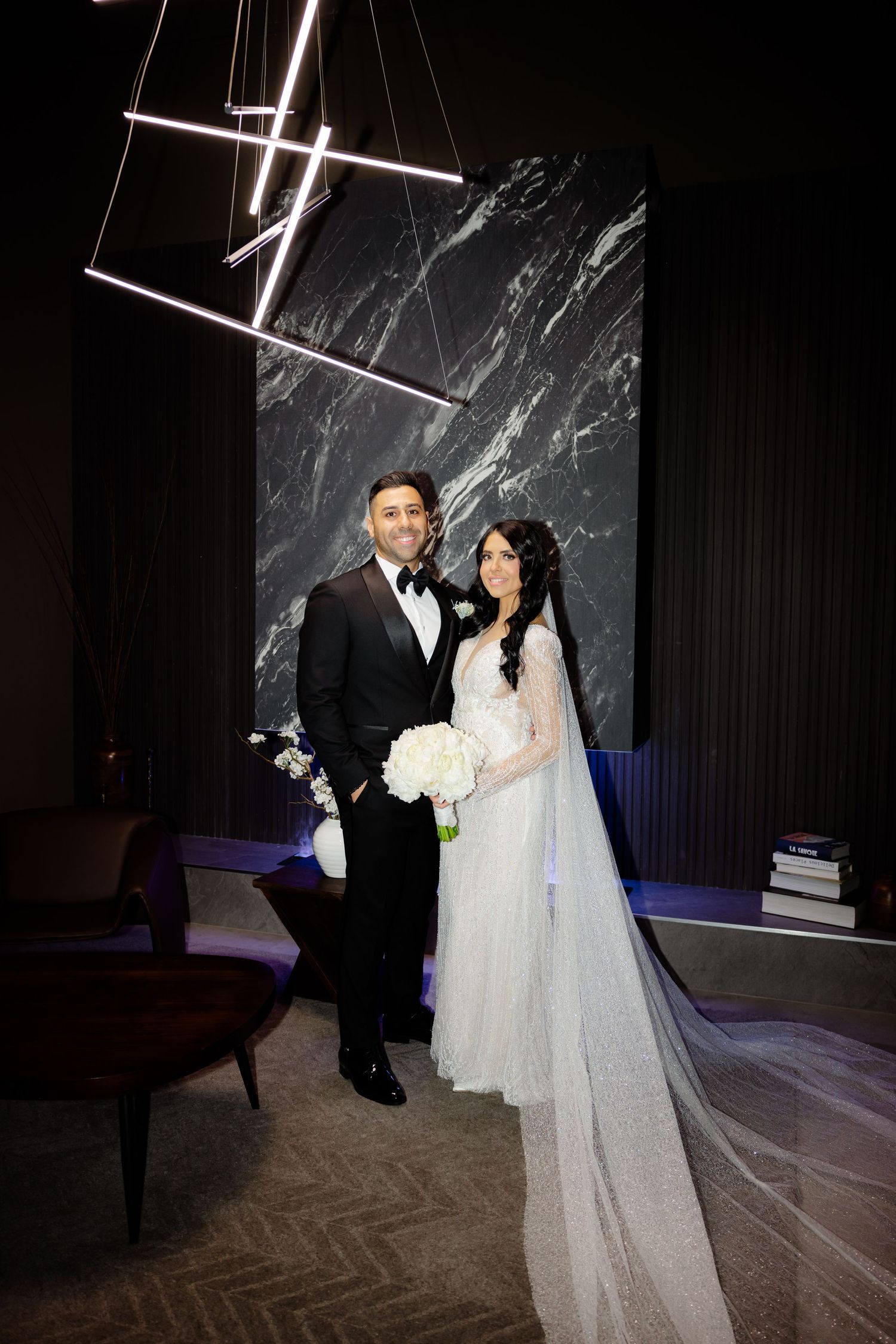 Wedding couple poses against modern dark marble wall with geometric light fixture overhead.